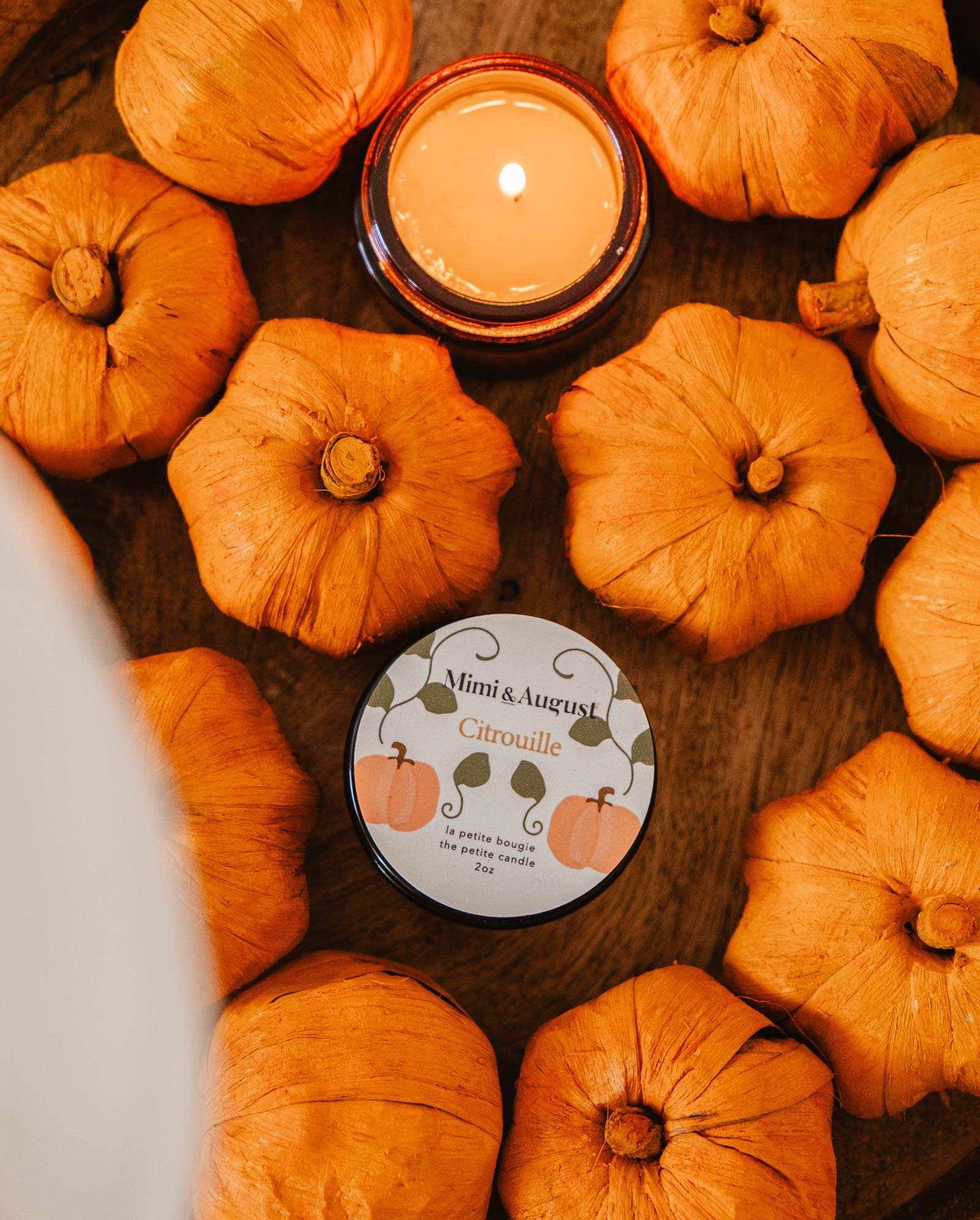 A wooden bowl featuring a lit Citrouille - Reusable Candle by Mimi & August, emitting a warm and soothing pumpkin fragrance, accompanied by faux orange pumpkins and a tin labeled "Mimi & August Citrouille.