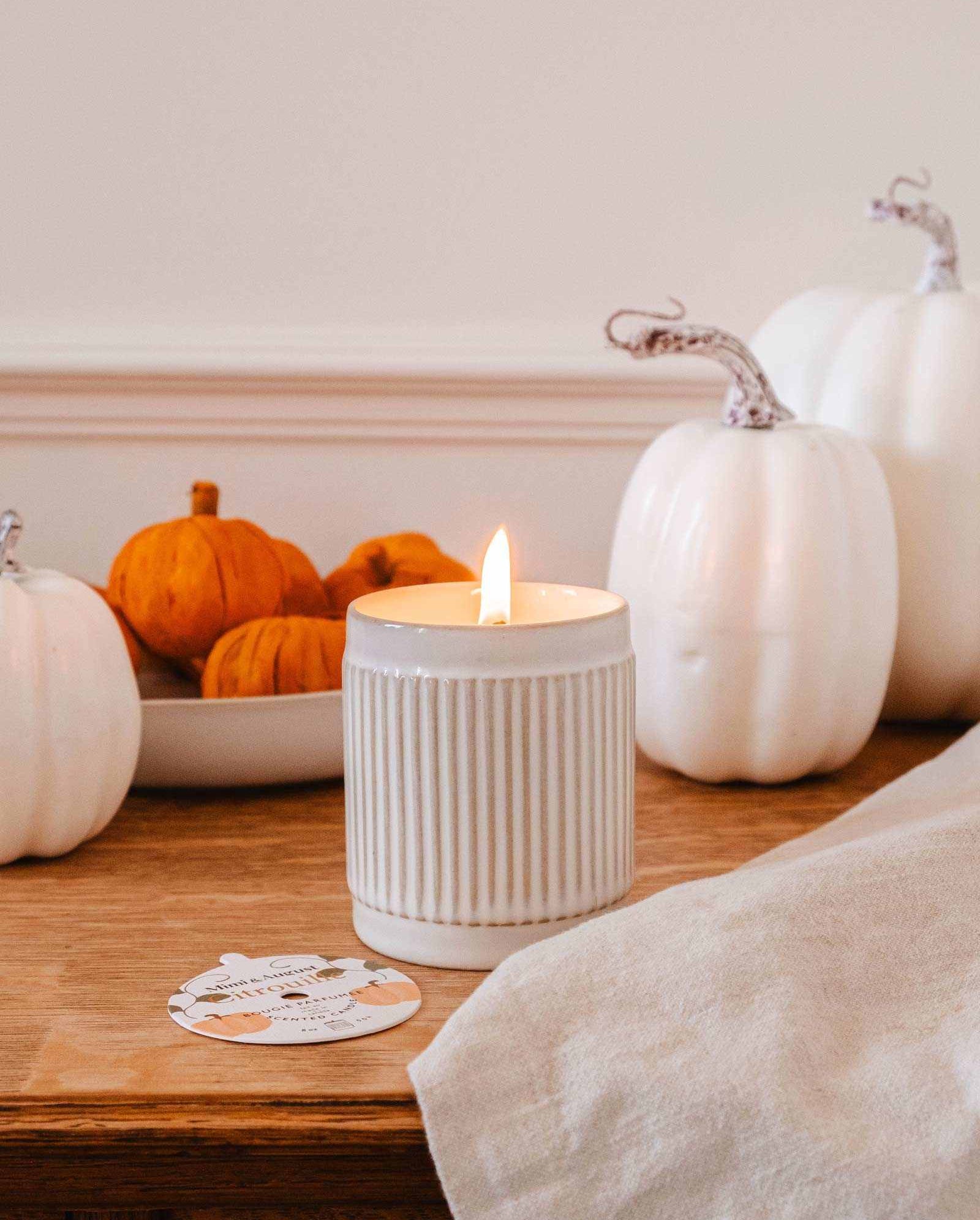 A lit Citrouille - Reusable Candle by Mimi & August is placed on a wooden table next to a cloth napkin, surrounded by white pumpkins and a bowl of small orange pumpkins, filling the air with a delightful pumpkin fragrance.