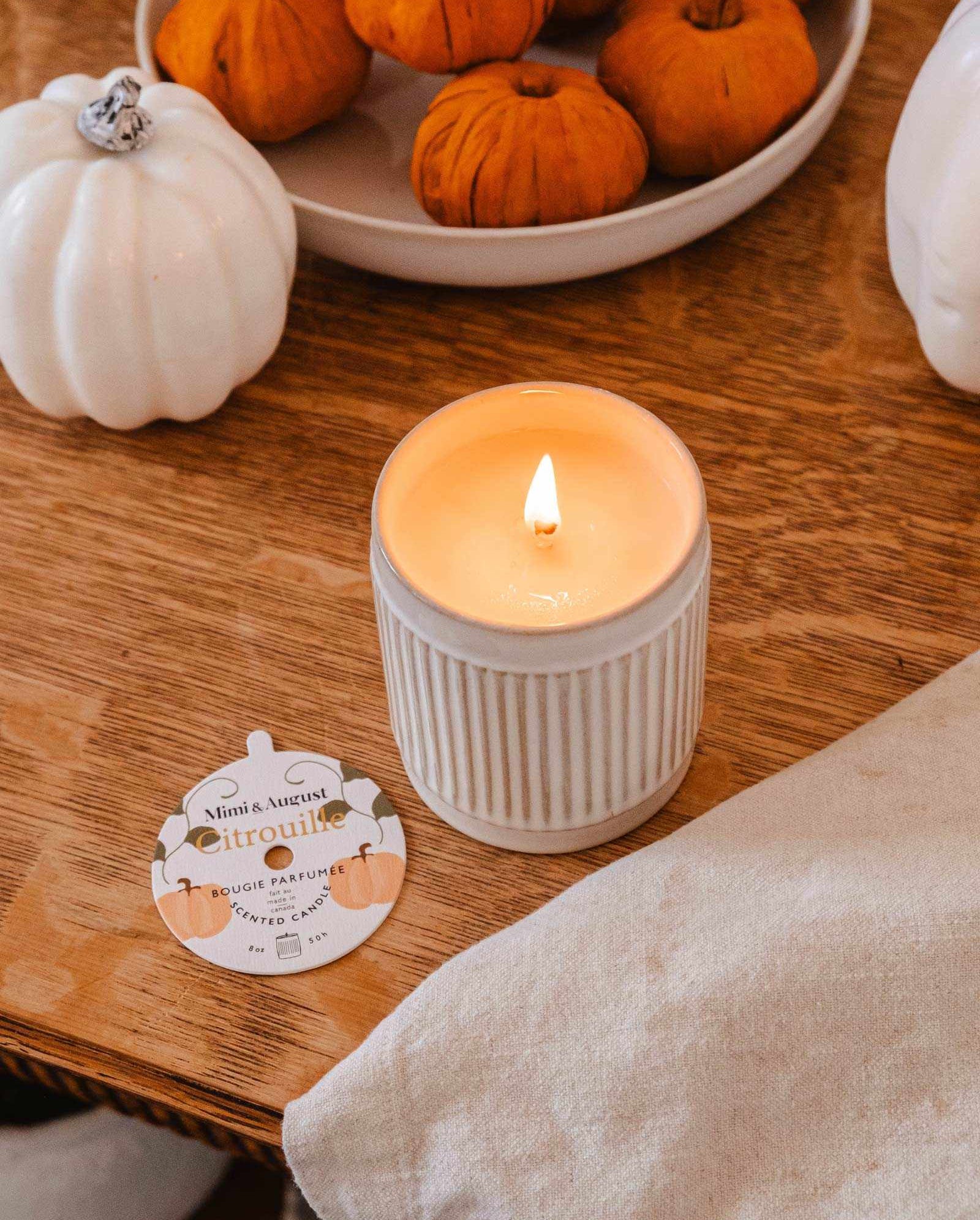 A lit **Citrouille - Reusable Candle** by **Mimi & August** sits on a wooden table next to a small dish of orange pumpkins and white pumpkin decorations. A card labeled "citrouille" is near the candle, emitting a delightful pumpkin fragrance.