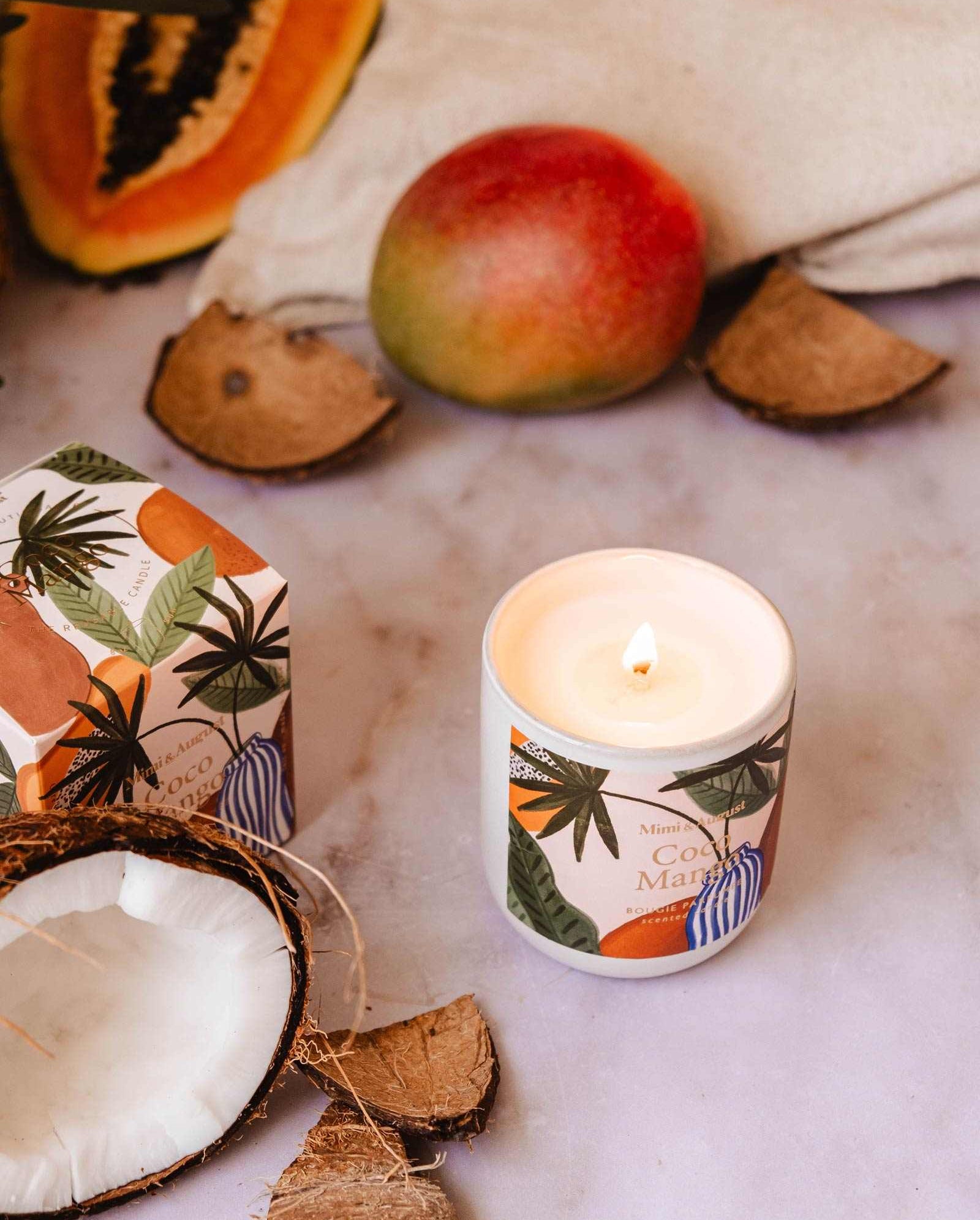 A Coco Mango - Reusable Candle by Mimi & August in a decorated container sits on a surface surrounded by coconut shells, a mango, a papaya slice, and a box with tropical designs, infusing the air with its tropical scent.