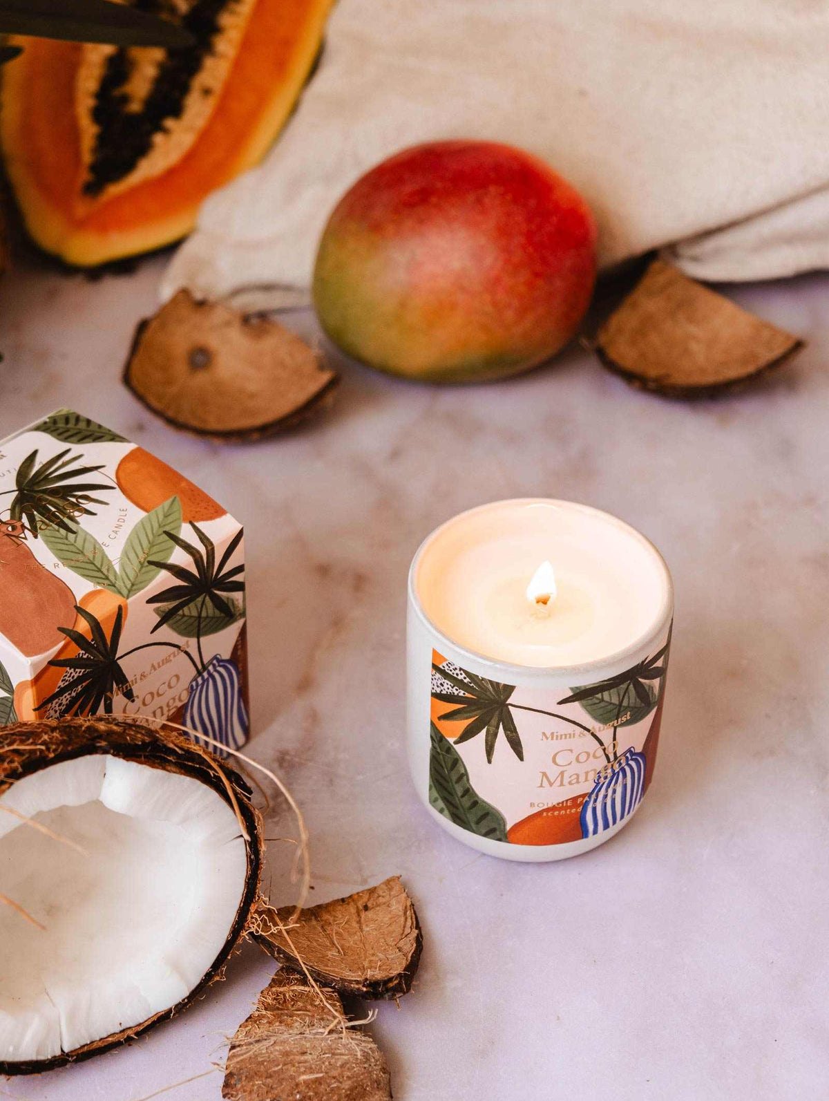 A Coco Mango - Reusable Candle by Mimi & August in a decorated container sits on a surface surrounded by coconut shells, a mango, a papaya slice, and a box with tropical designs, infusing the air with its tropical scent.