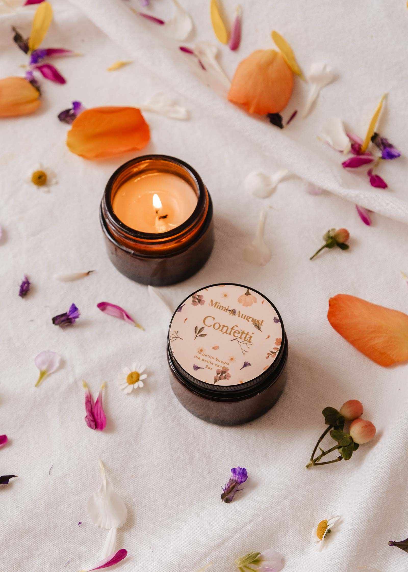 An open amber Mimi & August Confetti Reusable Soy Candle with a fruity mandarin scent glows beside its closed jar on a white cloth, surrounded by scattered flower petals.