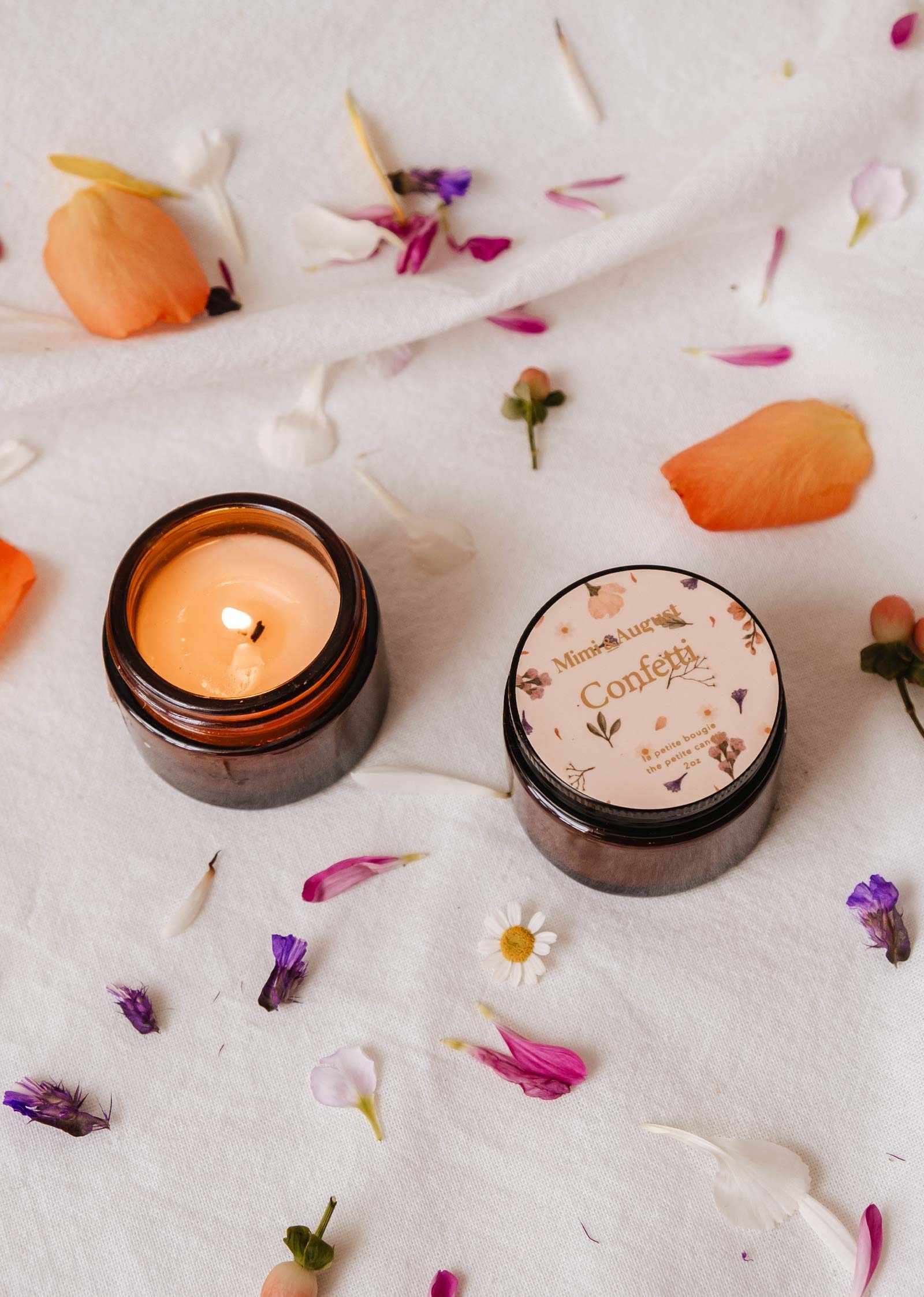 Two small amber glass jars from Mimi & August sit on white fabric with flower petals; one lit and fruity mandarin-scented, the other features a floral "Confetti - Reusable Soy Candle" label.