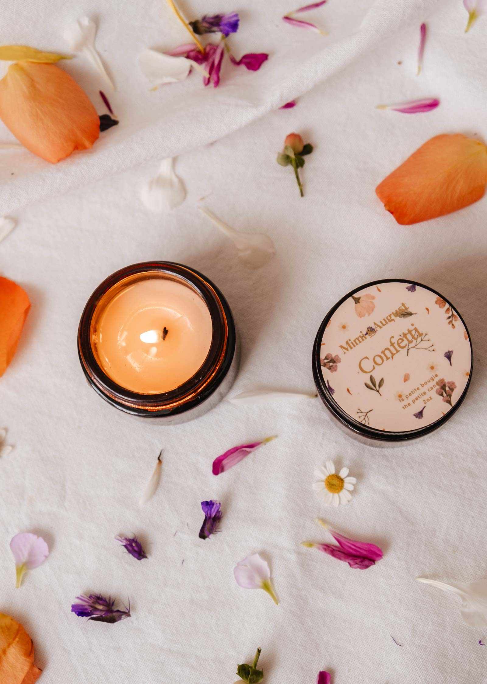 The Confetti - Reusable Soy Candle by Mimi & August, in a brown jar with a floral lid, sits on white cloth among flower petals and leaves, releasing a fruity mandarin aroma.