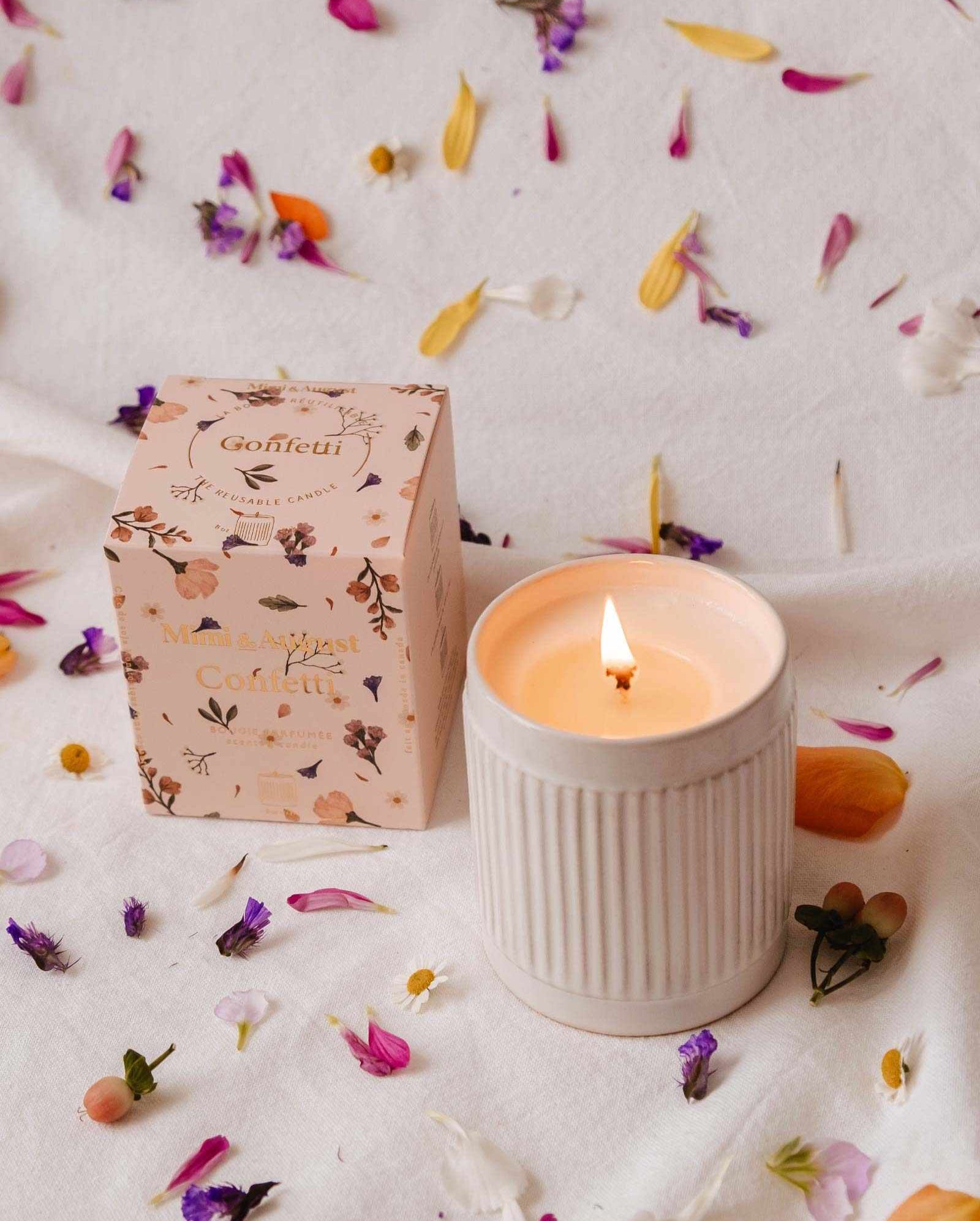 A lit Confetti - Reusable Soy Candle by Mimi & August in a ribbed ceramic holder sits beside a floral box, surrounded by colorful flower petals on a white surface, filling the air with fruity mandarin fragrance.