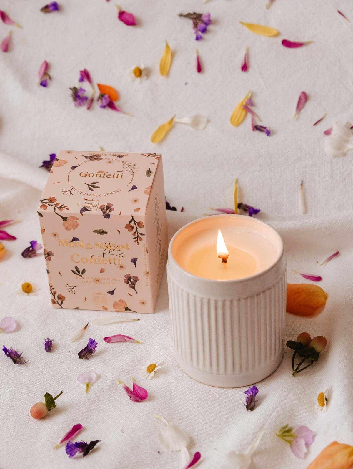 A lit Confetti - Reusable Soy Candle by Mimi & August in a ribbed ceramic holder sits beside a floral box, surrounded by colorful flower petals on a white surface, filling the air with fruity mandarin fragrance.