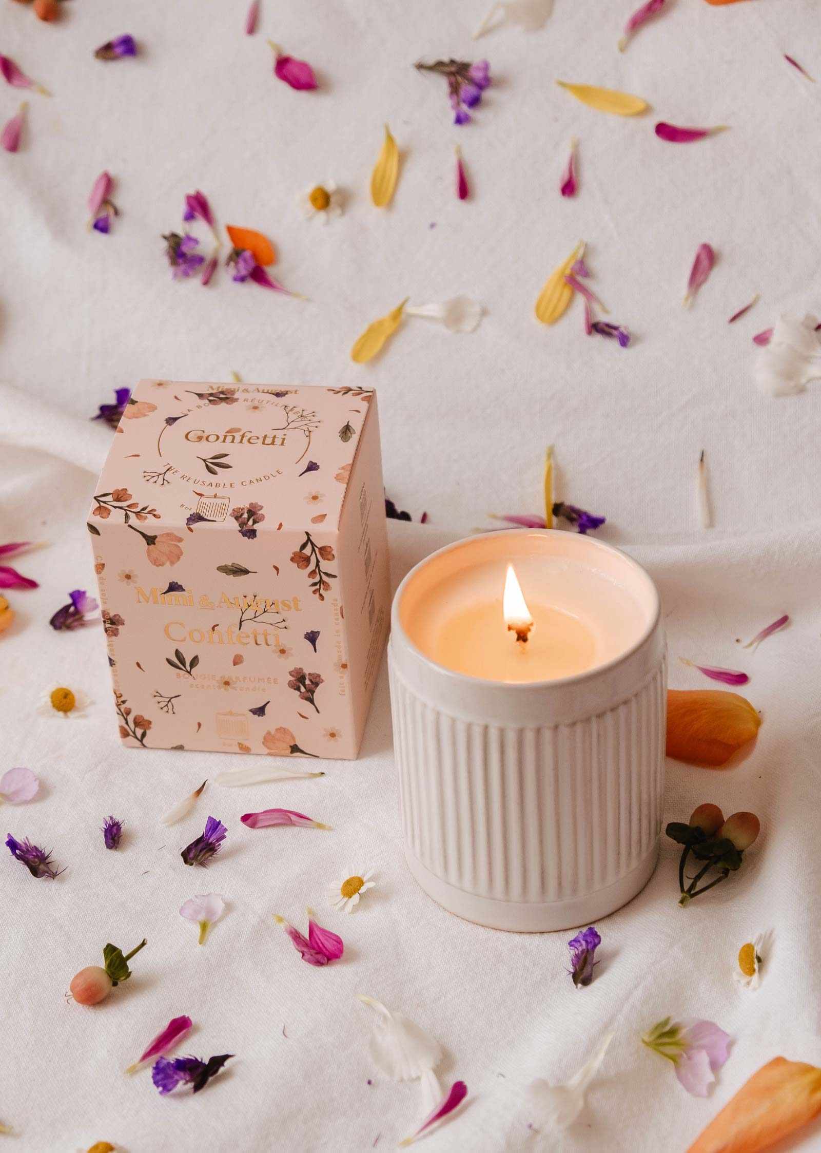Une bougie de soya réutilisable Confetti de Mimi & August allumée dans un bougeoir en céramique cannelé repose à côté de sa boîte florale, entourée de pétales de fleurs colorés sur une surface blanche, emplissant l'air d'un parfum fruité de mandarine.