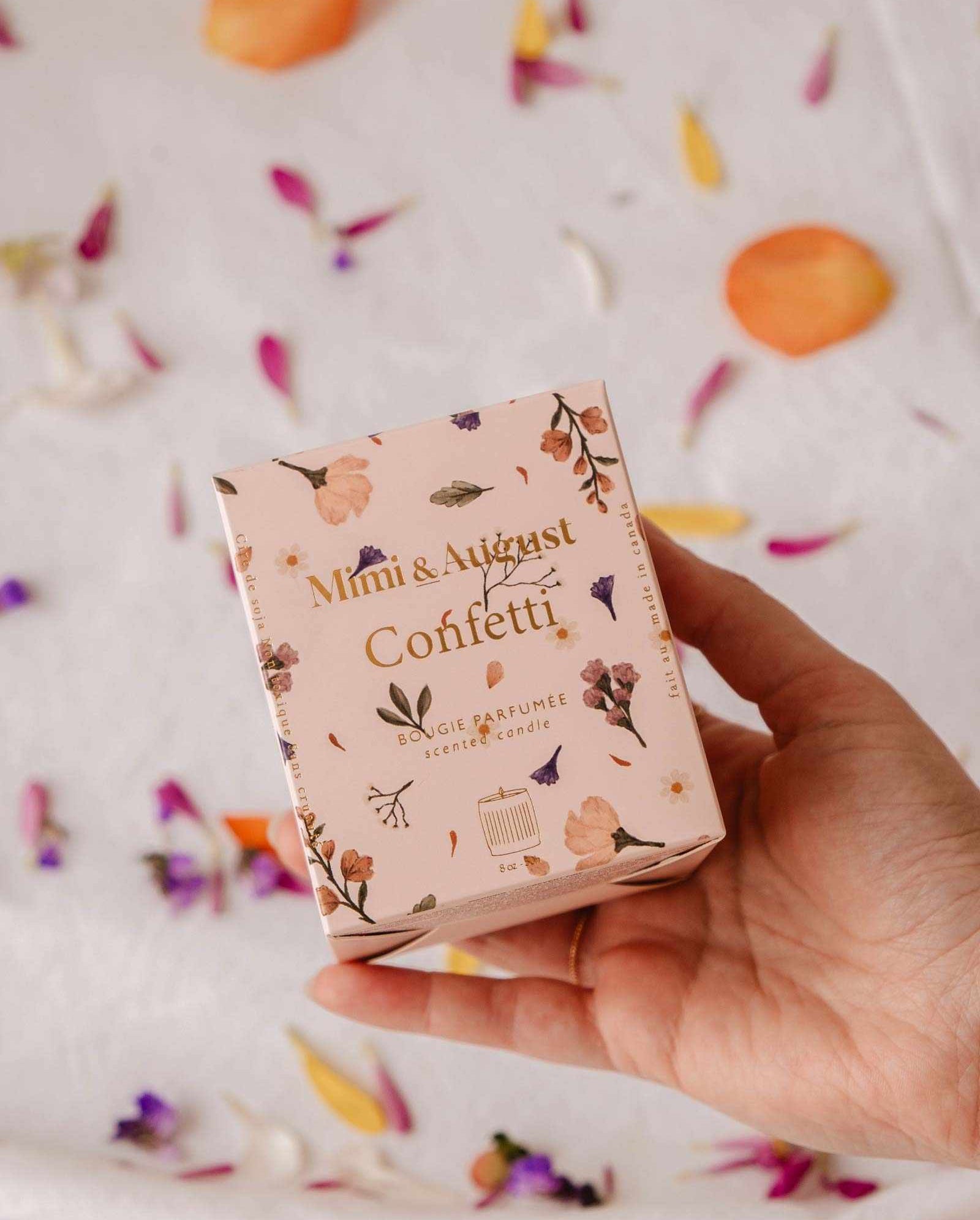 A hand holds a pink "Mimi & August Confetti - Reusable Soy Candle" box with floral designs; flower petals are scattered on the white surface in the background.