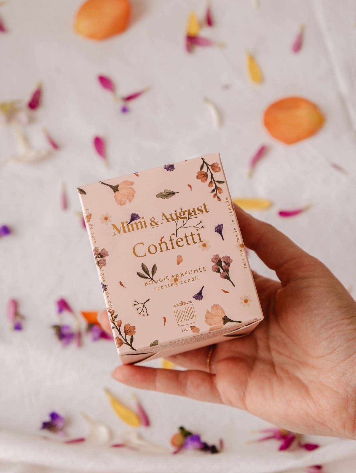 A hand holds a pink "Mimi & August Confetti - Reusable Soy Candle" box with floral designs; flower petals are scattered on the white surface in the background.