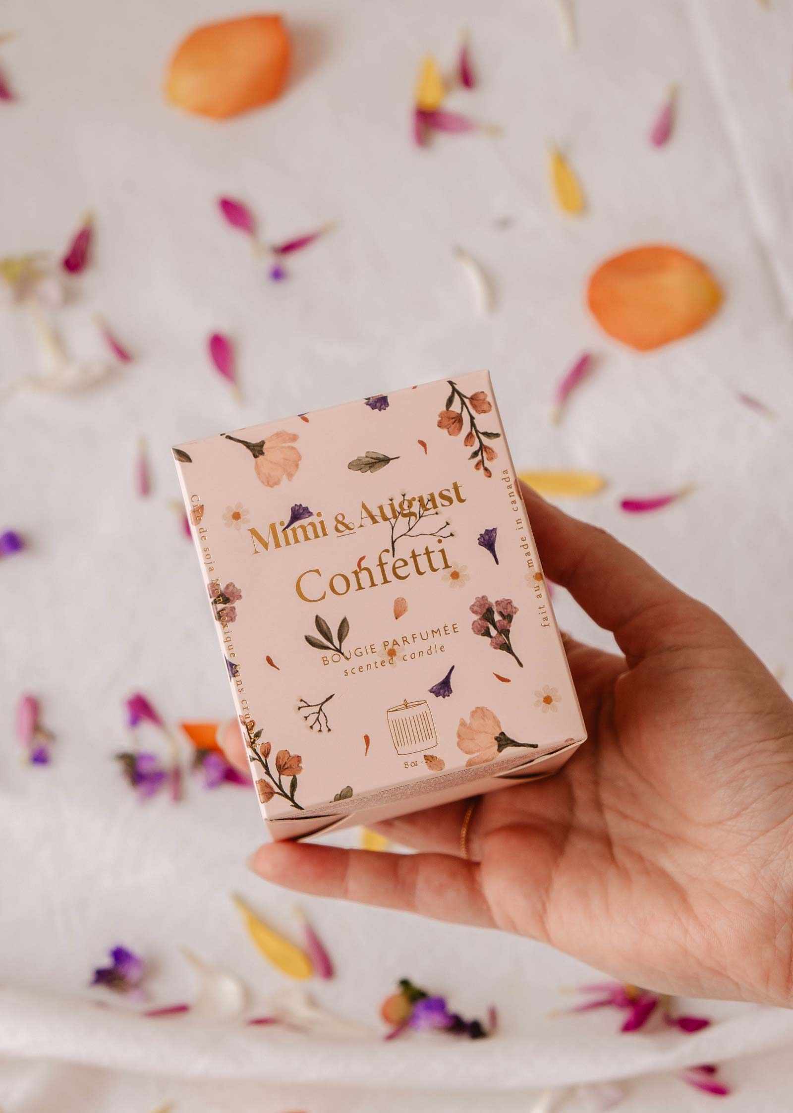 Une main tient une boîte rose « Mimi & August Confetti – Bougie de Soya Réutilisable » au design floral; des pétales de fleurs sont éparpillés sur la surface blanche en arrière-plan.