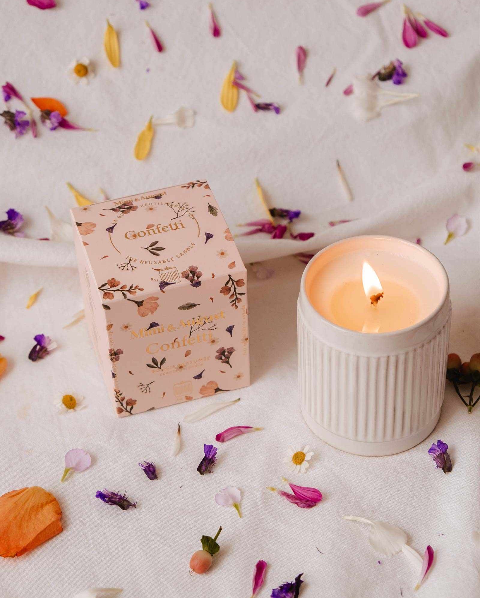 A lit Mimi & August Confetti - Reusable Soy Candle in a ribbed holder sits beside a floral box on a white cloth, surrounded by colorful petals. Enjoy its warm ambiance and subtle fruity mandarin scent.