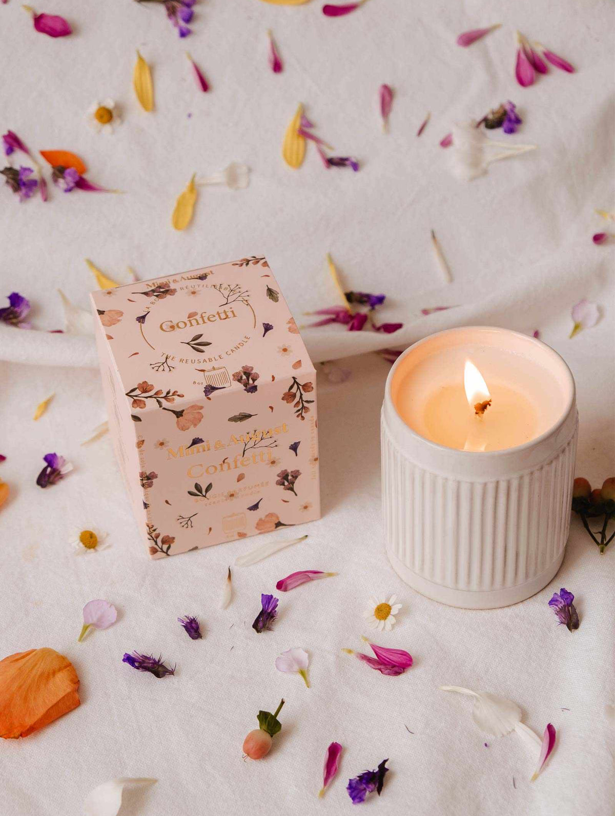 A lit Mimi & August Confetti - Reusable Soy Candle in a ribbed holder sits beside a floral box on a white cloth, surrounded by colorful petals. Enjoy its warm ambiance and subtle fruity mandarin scent.