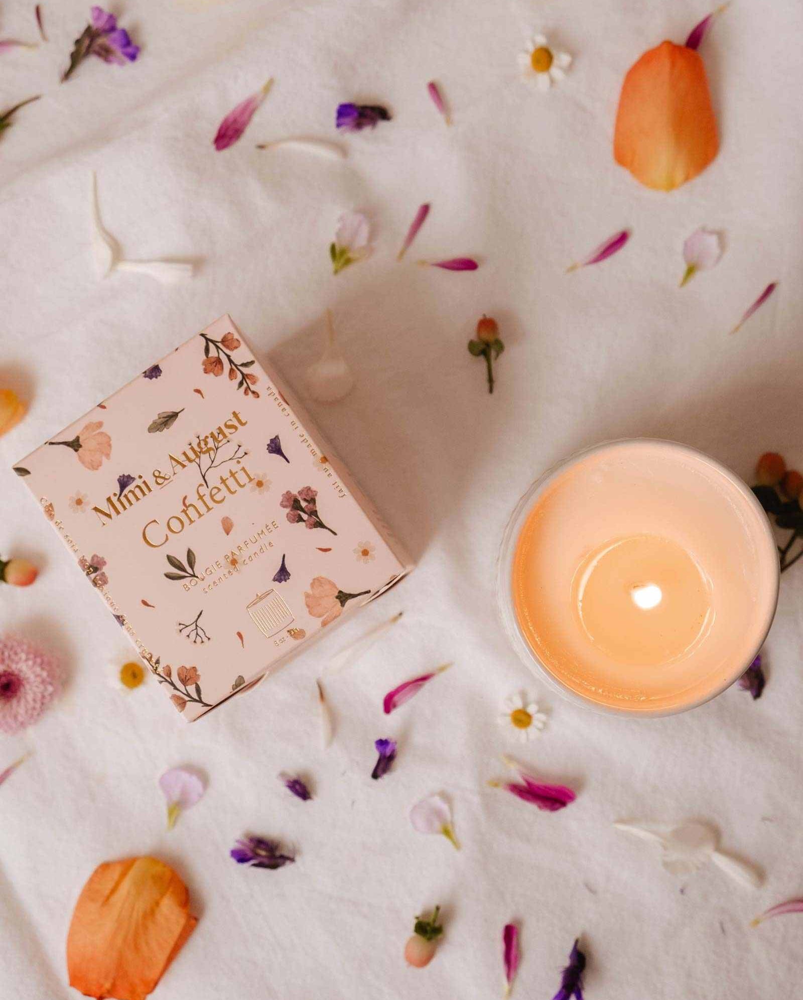 A lit Mimi & August Confetti - Reusable Soy Candle sits in a ceramic holder beside its floral box on white fabric strewn with colorful petals, evoking a festive atmosphere with a touch of fruity mandarin scent.