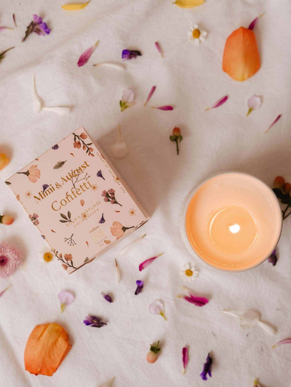 A lit Mimi & August Confetti - Reusable Soy Candle sits in a ceramic holder beside its floral box on white fabric strewn with colorful petals, evoking a festive atmosphere with a touch of fruity mandarin scent.