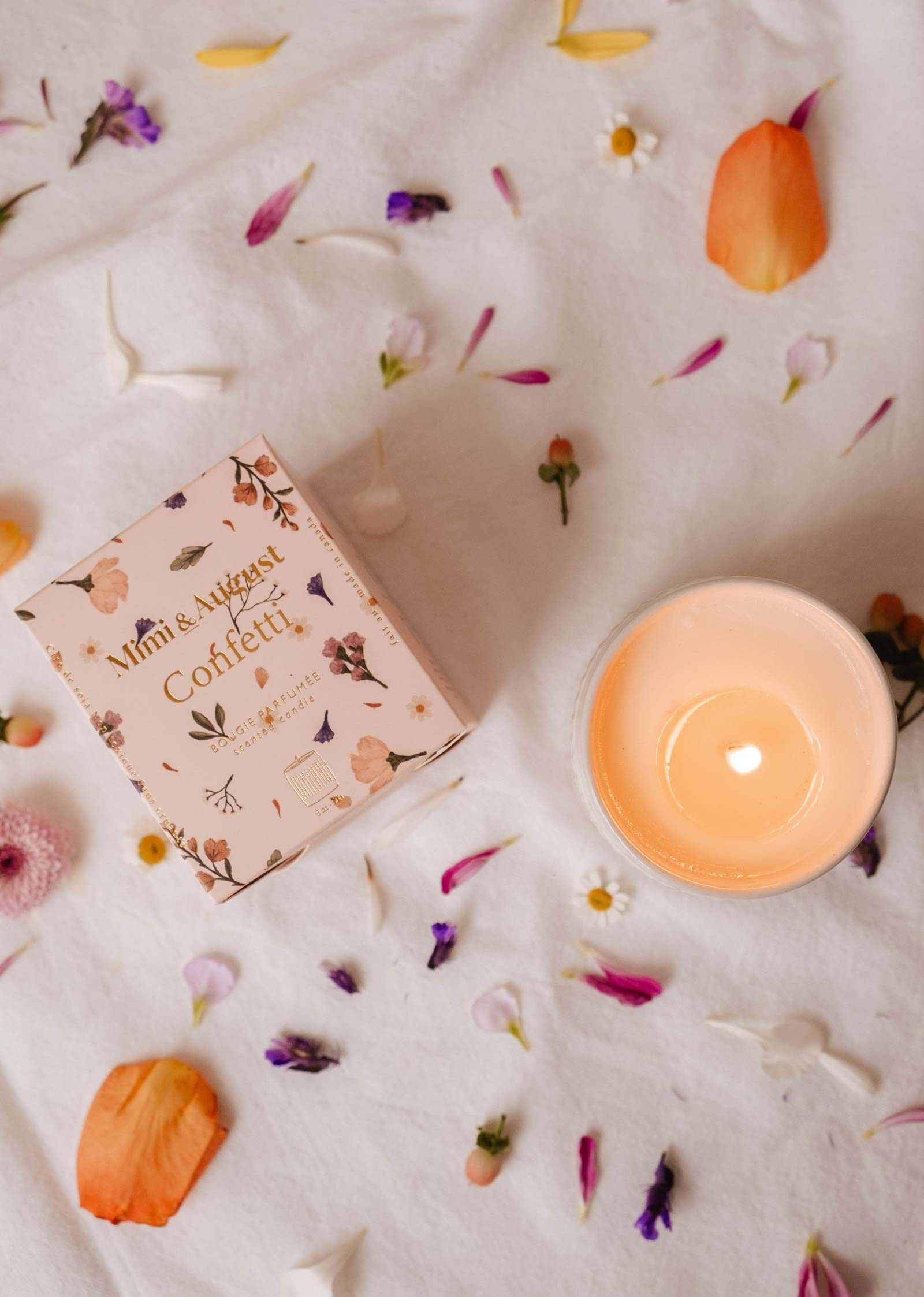 Une bougie de soya réutilisable Confetti de Mimi & August allumée repose dans un bougeoir en céramique à côté de sa boîte florale sur un tissu blanc parsemé de pétales colorés, évoquant une atmosphère festive avec une touche de mandarine fruitée.