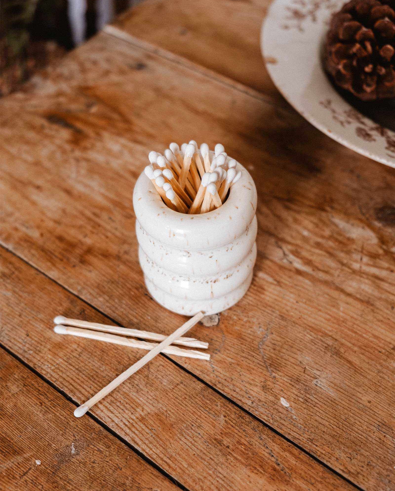 The Mimi & August Ceramic Match Pot, filled with cotton swabs, sits on a wooden table with four swabs beside it—perfect as a candle accessory for any space.