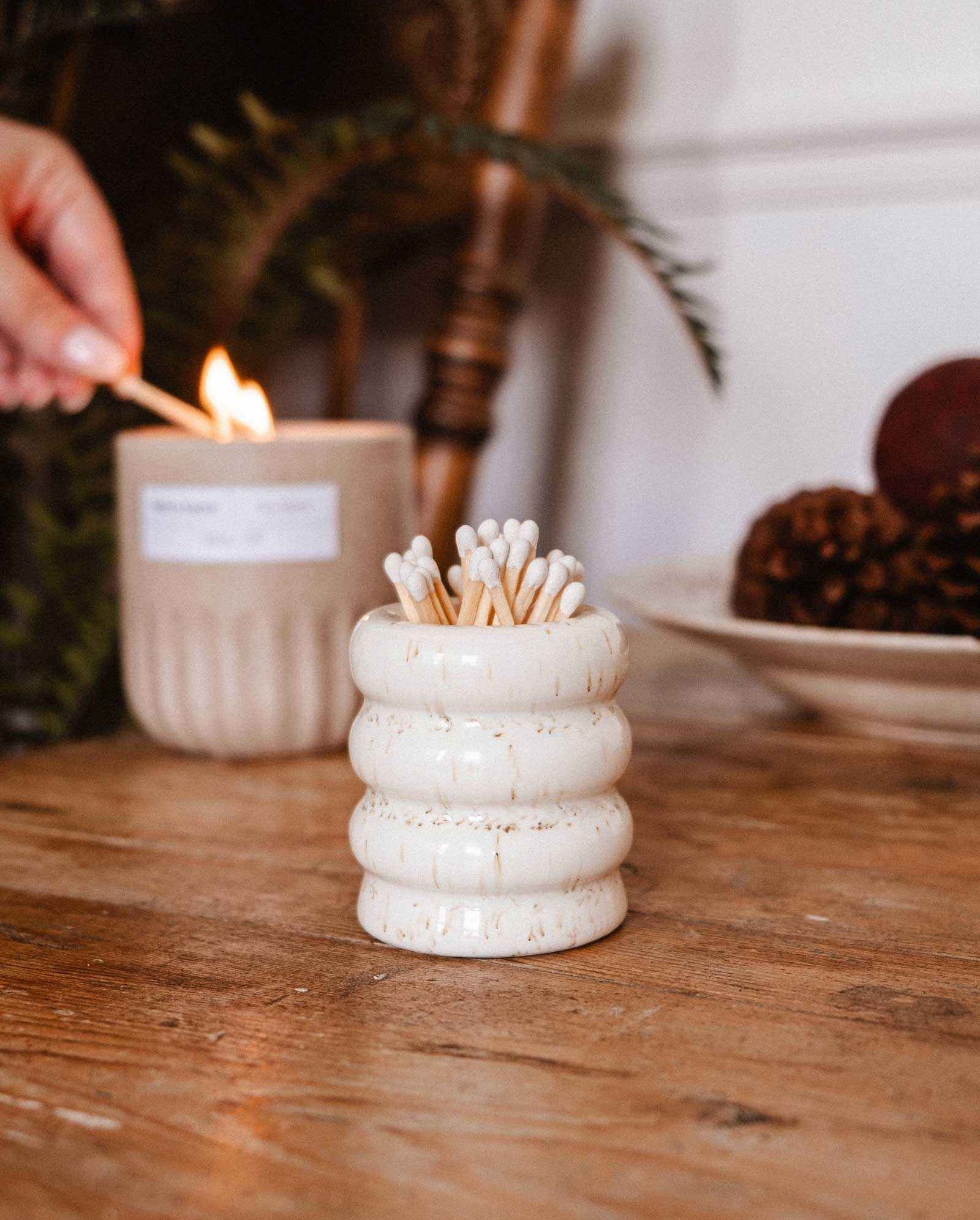 The Mimi & August Ceramic Match Pot, filled with wooden matches, sits on a wooden table as a hand lights a candle in the background, showcasing stylish candle accessories.