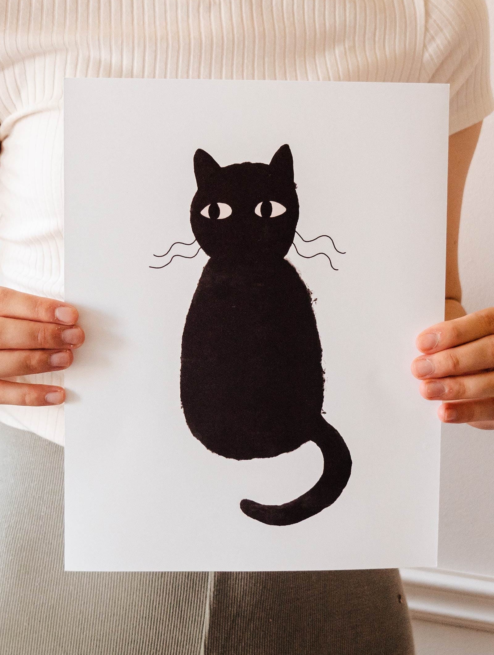 A woman displaying a Cute Black Cat Art Print by Mimi & August on recycled paper.