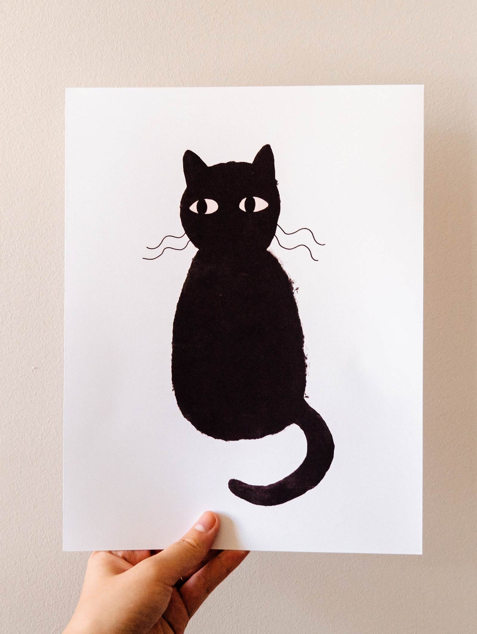 A person showcasing a Cute Black Cat Art Print by Mimi & August on recycled paper.
