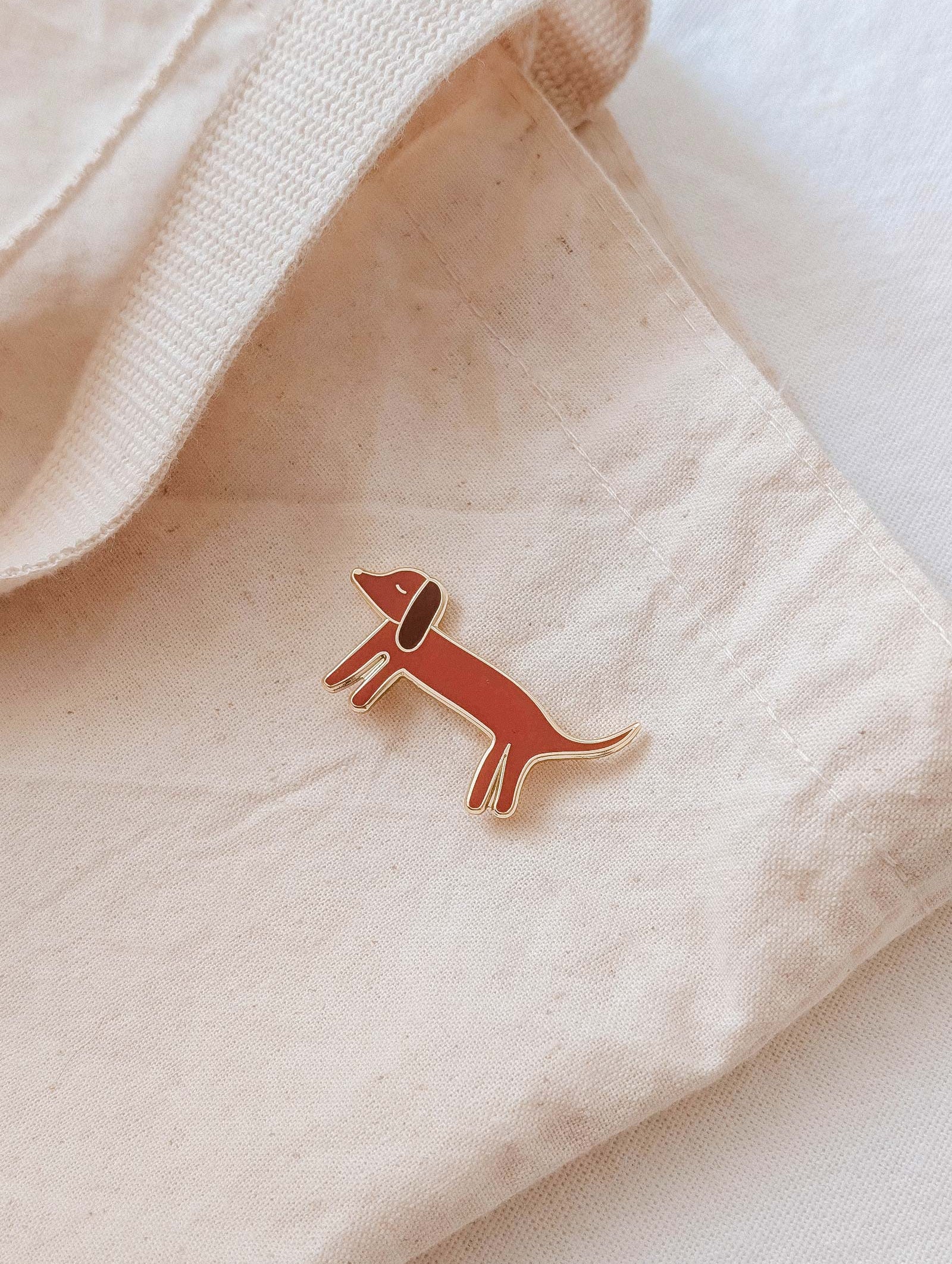 The Mimi & August Dachshund Enamel Pin, shaped like a wiener dog, is attached to a beige fabric tote bag—an adorable accessory for any dog lover.