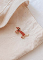 The Mimi & August Dachshund Enamel Pin, shaped like a wiener dog, is attached to a beige fabric tote bag—an adorable accessory for any dog lover.