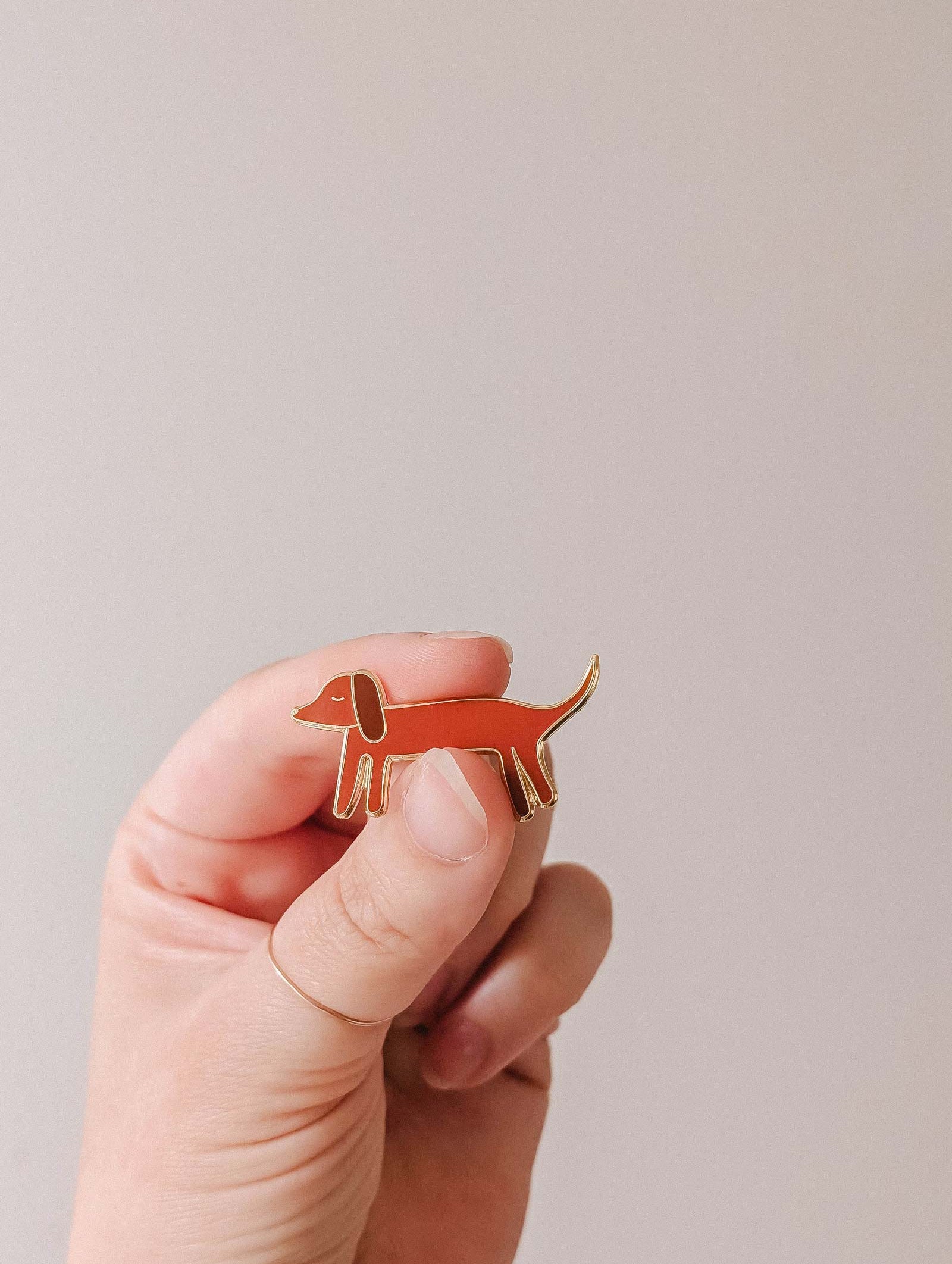 A hand holds the Mimi & August Dachshund Enamel Pin, shaped like a brown wiener dog—perfect for dog lovers—against a plain light background.