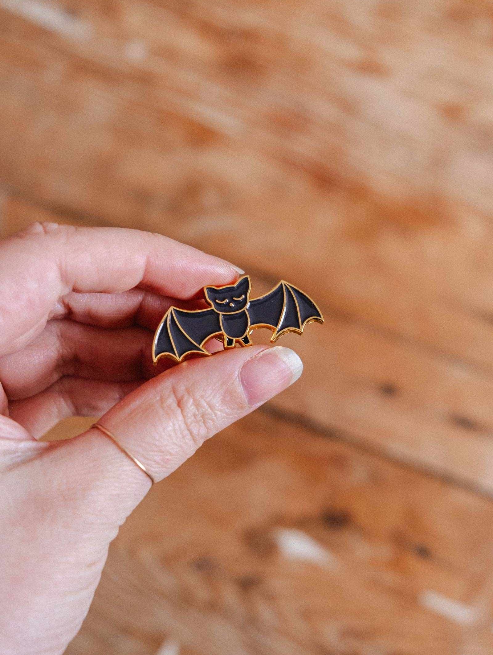 A hand displays the Mimi & August Little Bat Enamel Pin, a cute black bat with gold outlines—an ideal gift for any animal lover.