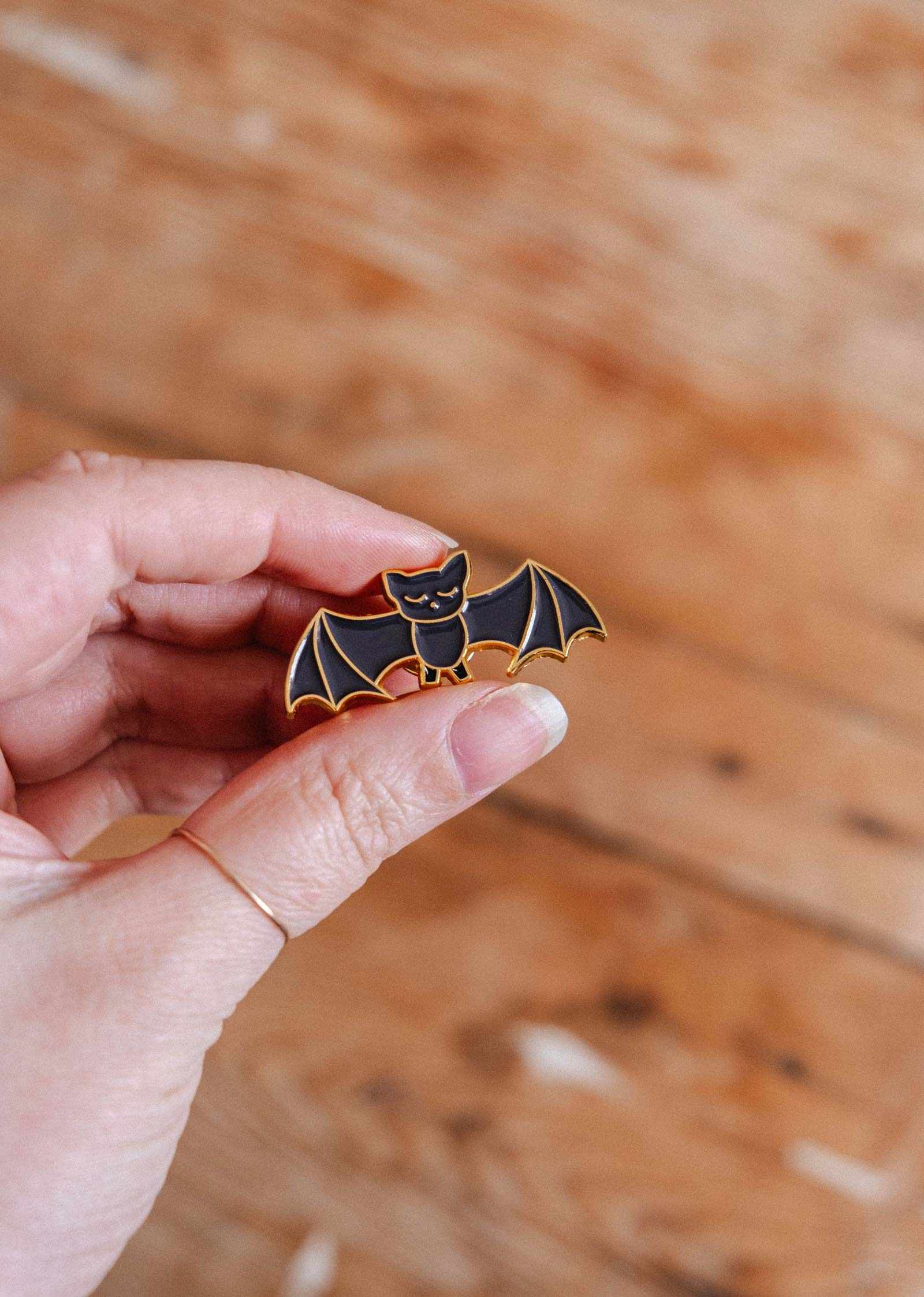 A hand displays the Mimi & August Little Bat Enamel Pin, a cute black bat with gold outlines—an ideal gift for any animal lover.