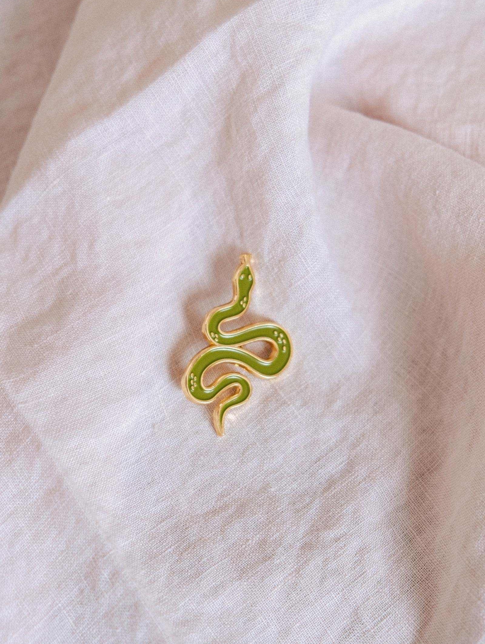 The Mimi & August Cute Serpent Enamel Pin in gold and green rests on a textured white fabric—an ideal gift for animal lovers.
