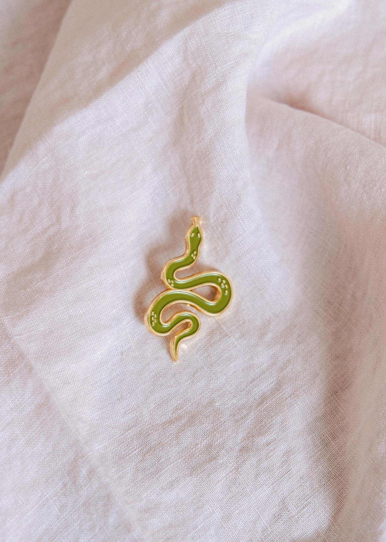 The Mimi & August Cute Serpent Enamel Pin in gold and green rests on a textured white fabric—an ideal gift for animal lovers.