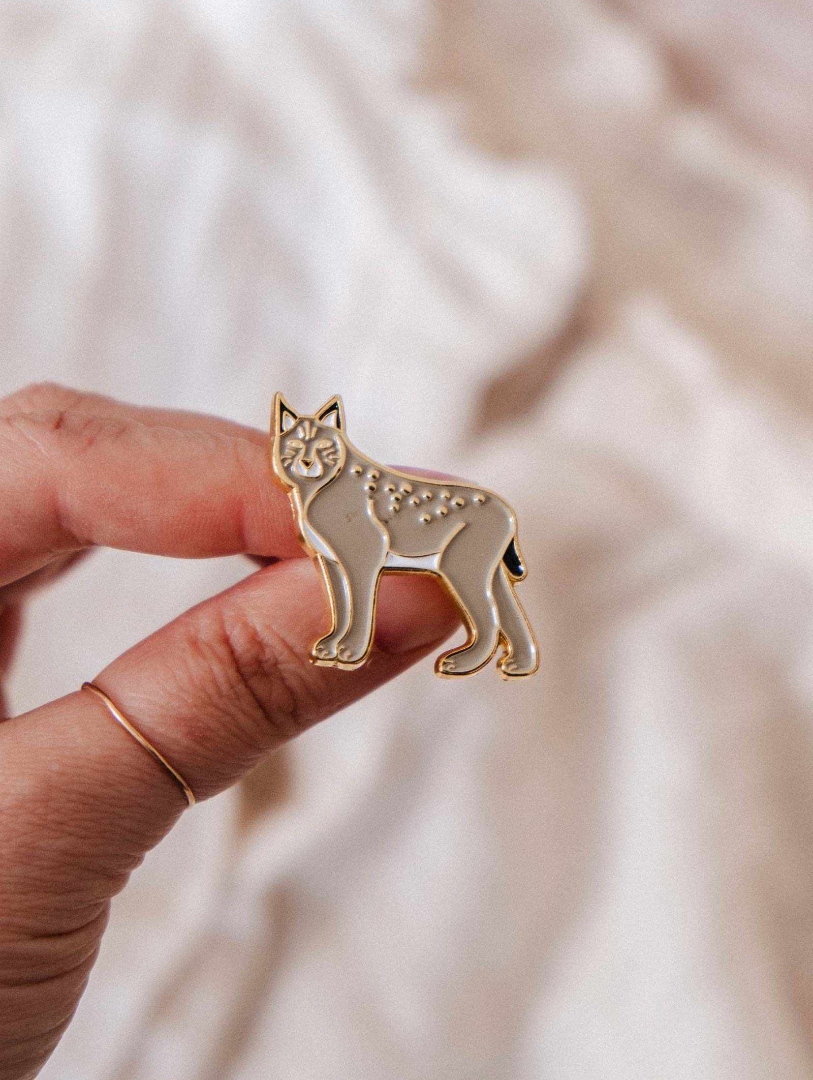 A hand displays the Lynx Enamel Pin by Mimi & August, with gold outlines and neutral tones—ideal for wildlife lovers—set against a soft, light-colored background.