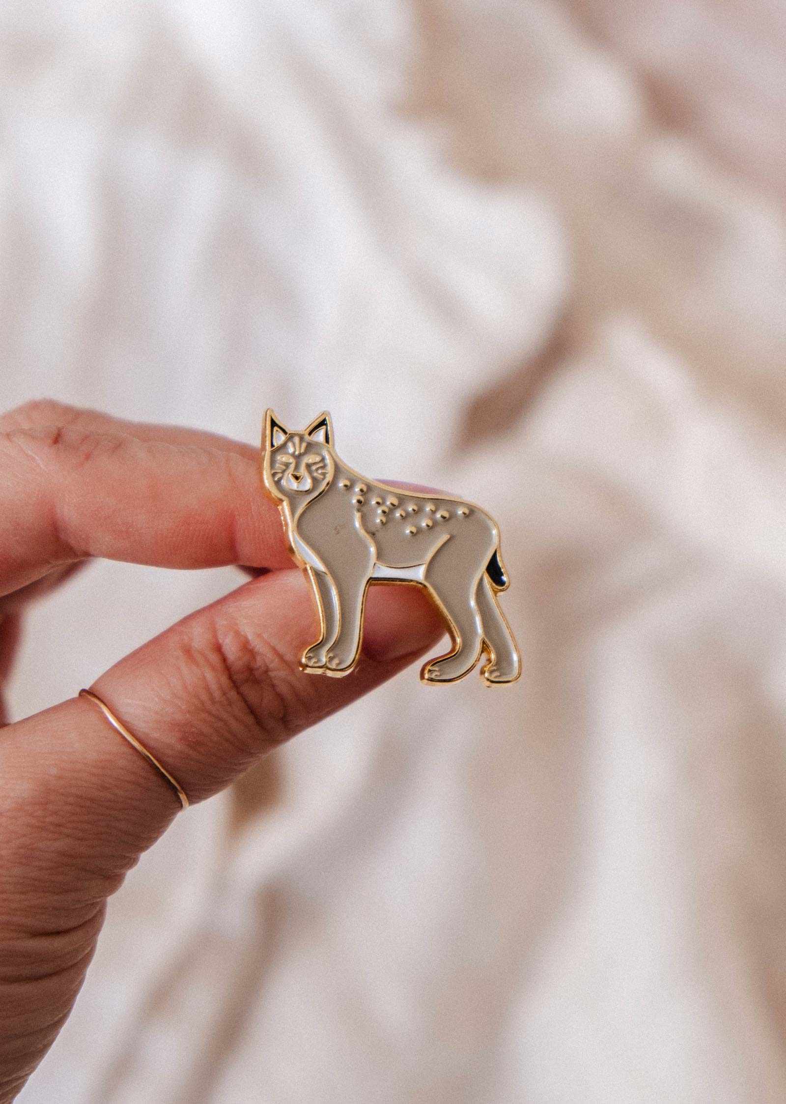 A hand displays the Lynx Enamel Pin by Mimi & August, with gold outlines and neutral tones—ideal for wildlife lovers—set against a soft, light-colored background.