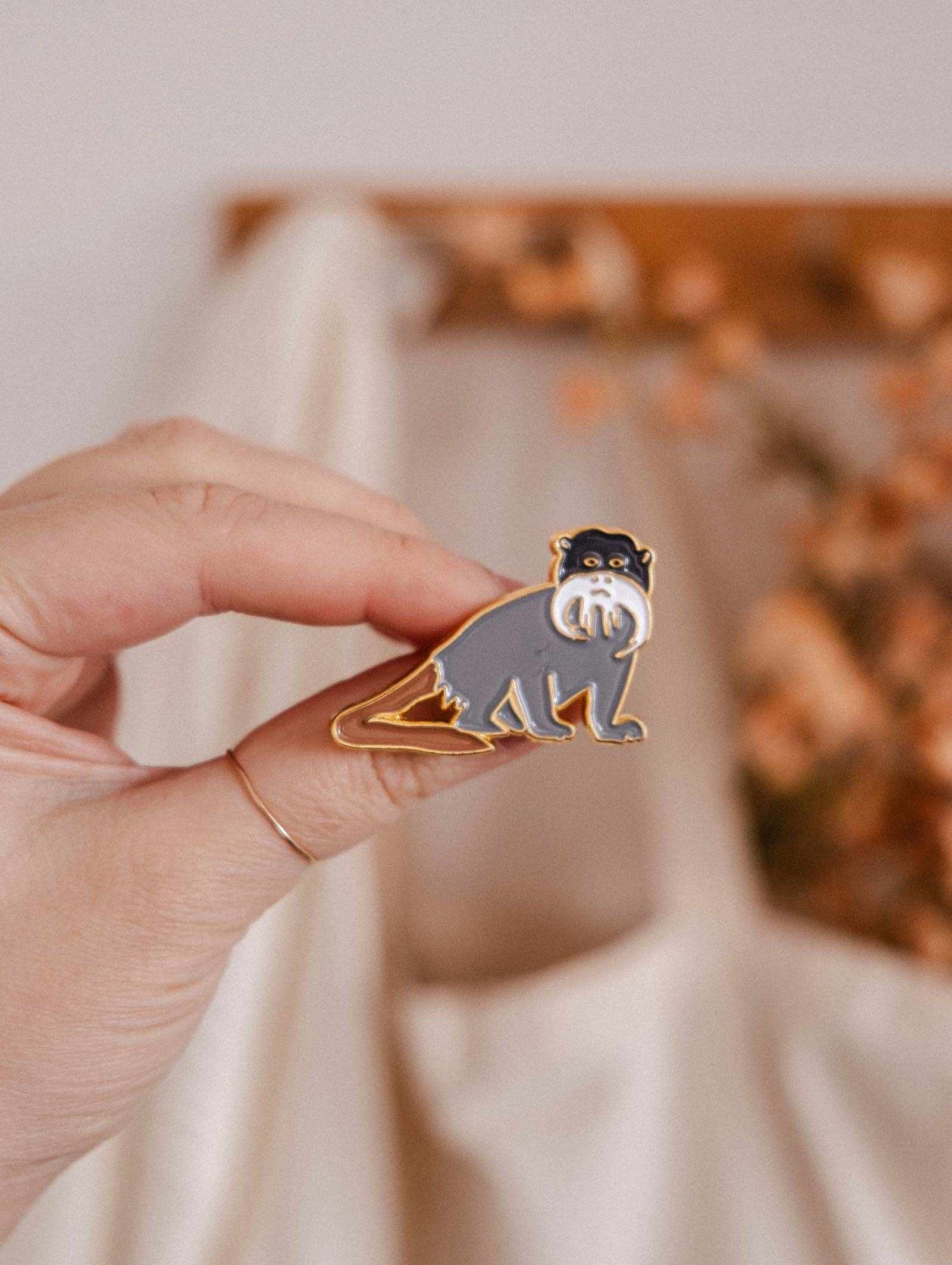 A hand displays the Sir Moustache Enamel Pin by Mimi & August—a charming gray and white monkey with a curled tail—against a softly blurred beige and brown background, ideal for animal lovers.