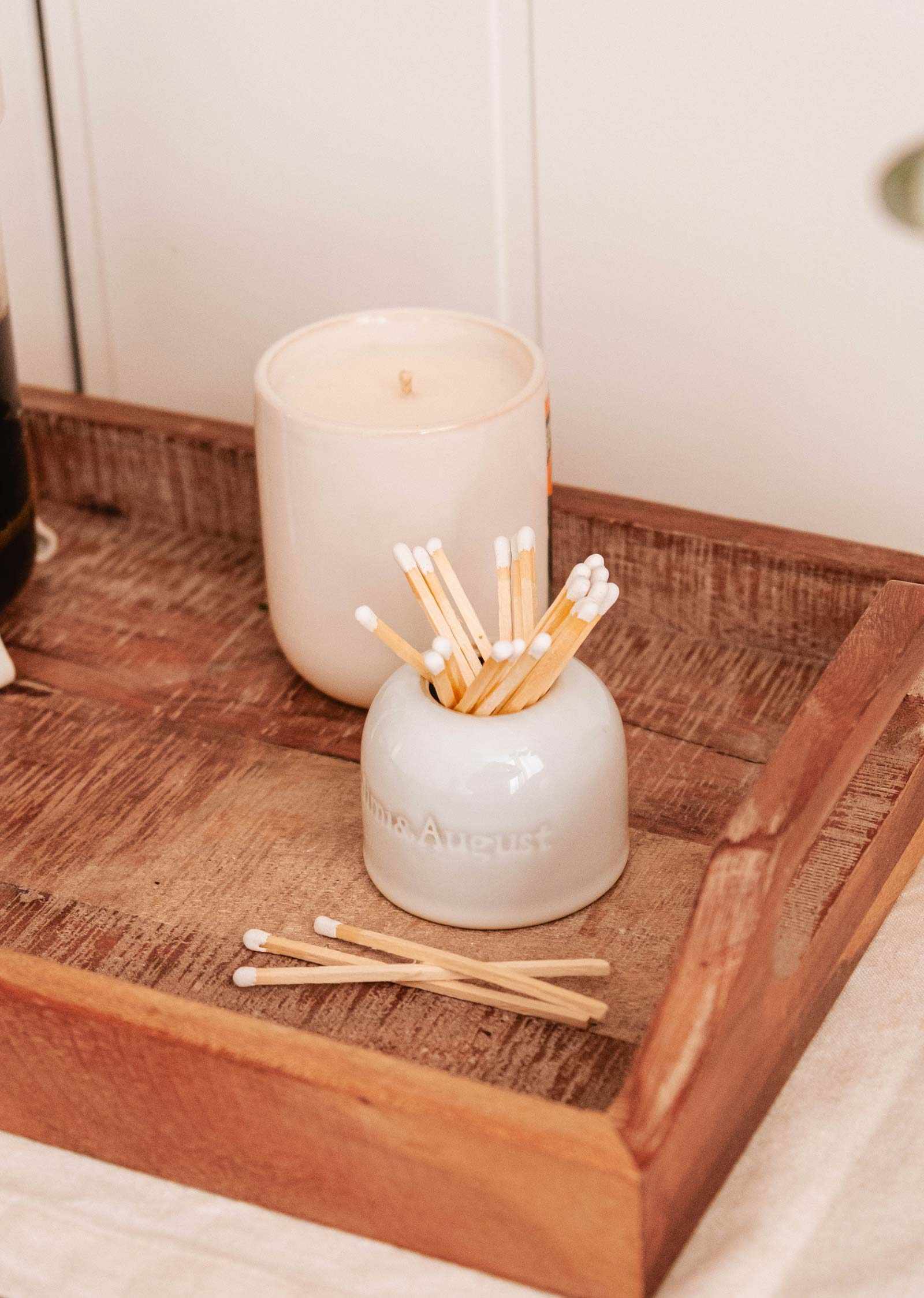 The Étincelles Ceramic Match Holder by Mimi & August sits on a wooden tray with a lit candle and serves as a multifunctional holder for cotton swabs, some of which are scattered artfully around.
