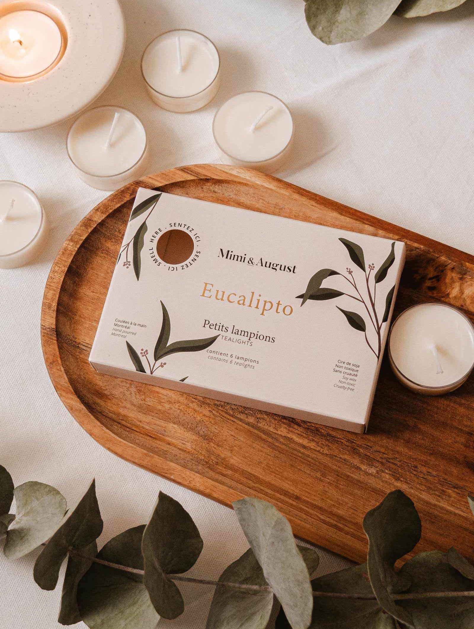 The Eucalipto Tealight Candle Set by Mimi & August is displayed on a wooden tray, surrounded by lit candles and eucalyptus leaves, creating a soothing atmosphere.