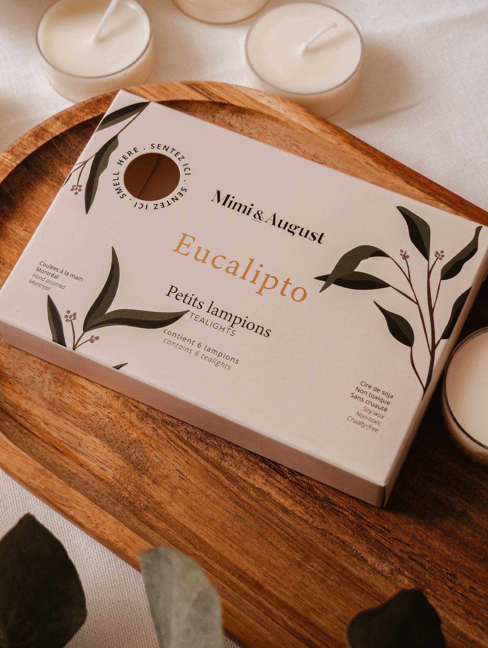 The Eucalipto Tealight Candle Set by Mimi & August sits elegantly on a wooden tray, surrounded by loose tea lights and green leaves, evoking a calming spa atmosphere.