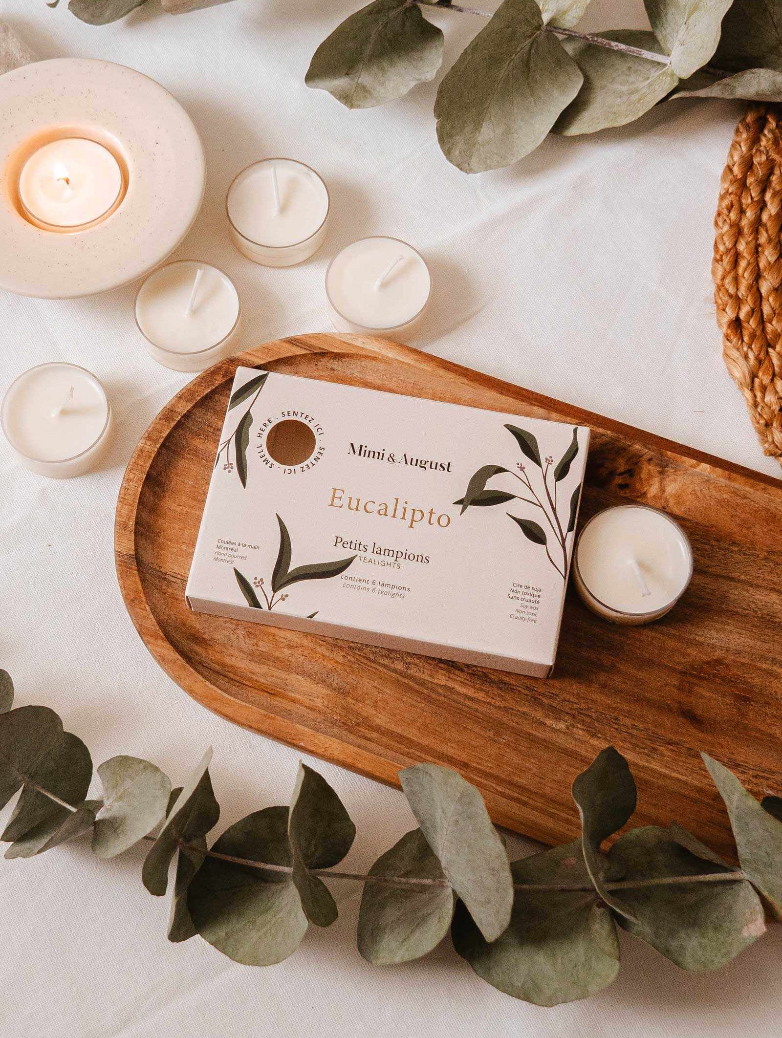 The "Eucalipto Tealight Candle Set" by Mimi & August rests on a wooden tray with eucalyptus leaves and lit candles, evoking a spa-like atmosphere that invites relaxation.