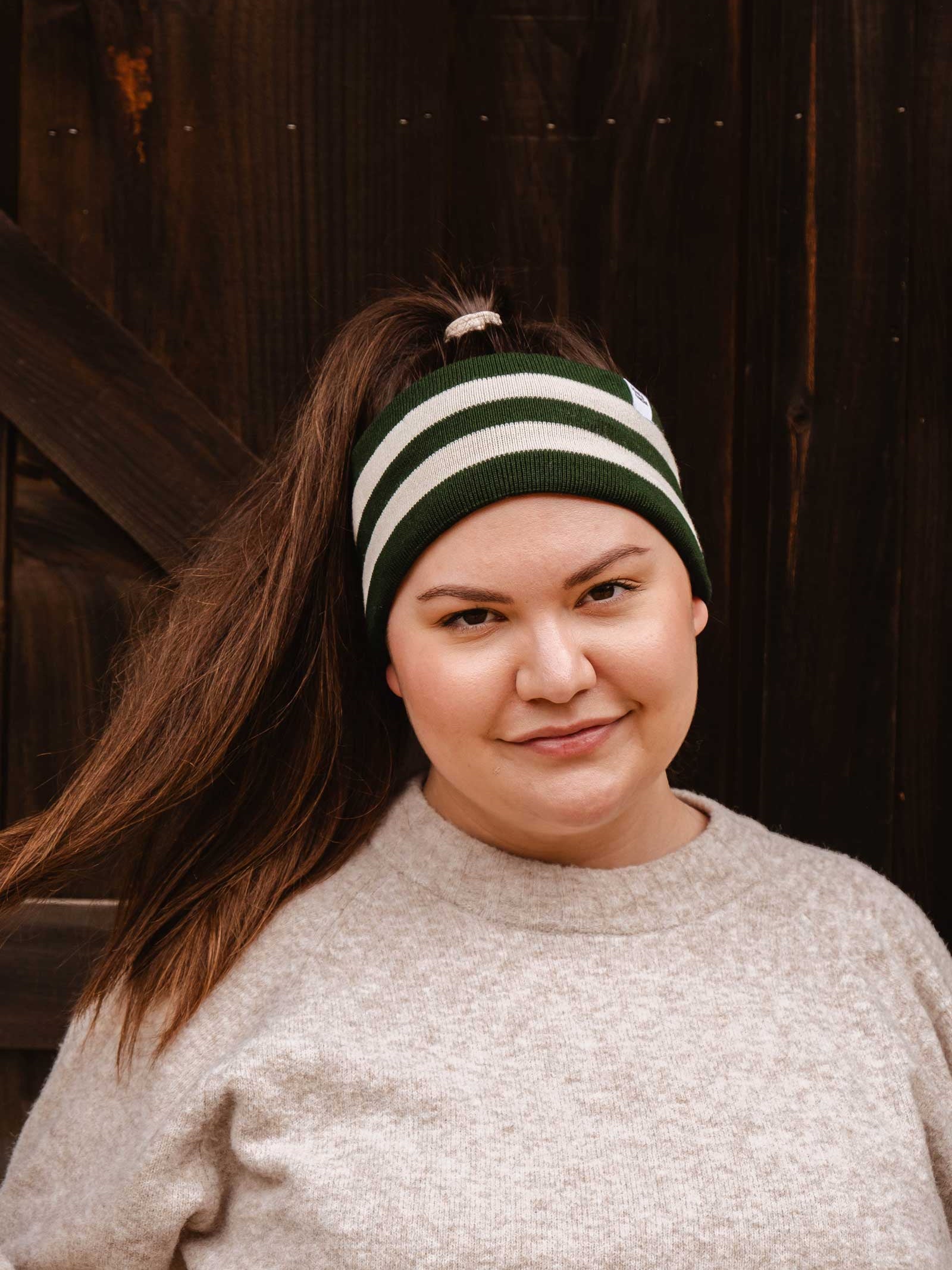 A person with long brown hair in a high ponytail, wearing the Mimi & August Evergreen Stripes Headband and a beige sweater featuring soft black fleece lining, stands in front of a dark wooden background.