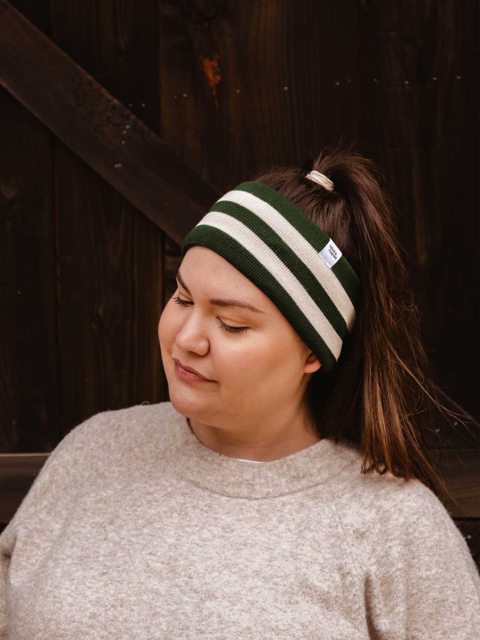 A person with long hair in a ponytail stands against a dark wooden background, wearing the Mimi & August Evergreen Stripes Headband—a green and white unisex acrylic knit with a cozy fleece lining—and a light grey sweater.