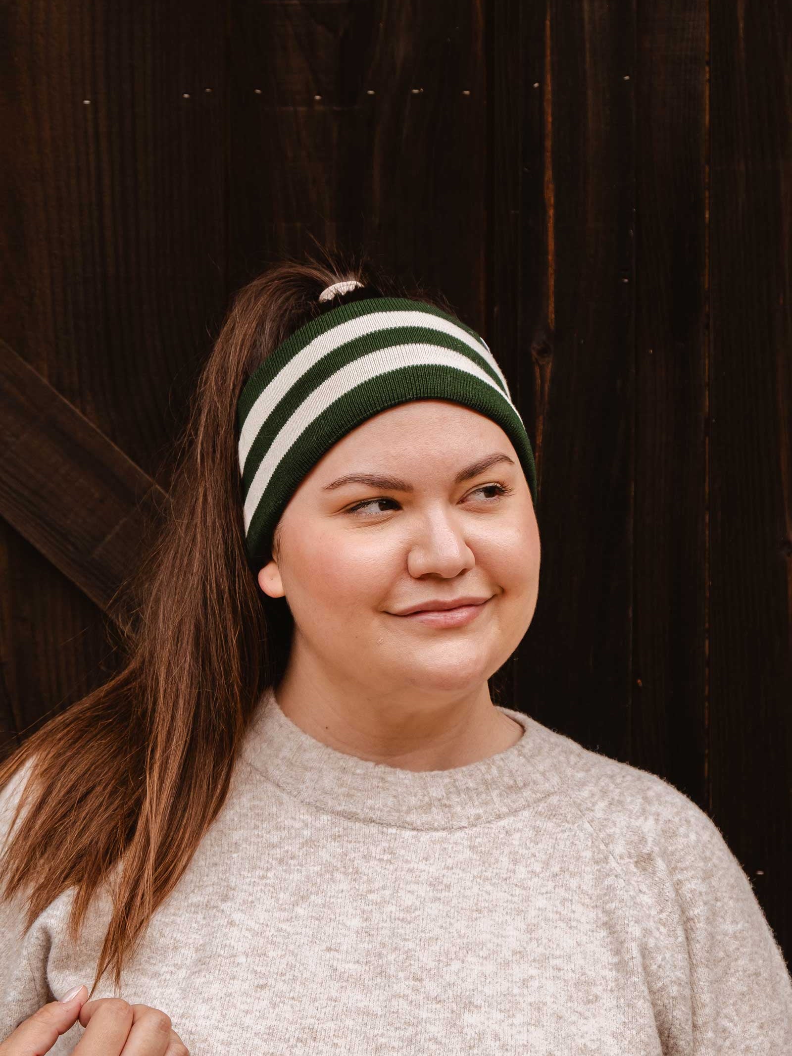 A person wearing a Mimi & August Evergreen Stripes Headband and a gray sweater looks to the side while standing in front of a dark wooden background. The unisex acrylic knit headband features green and white stripes, providing both style and warmth.