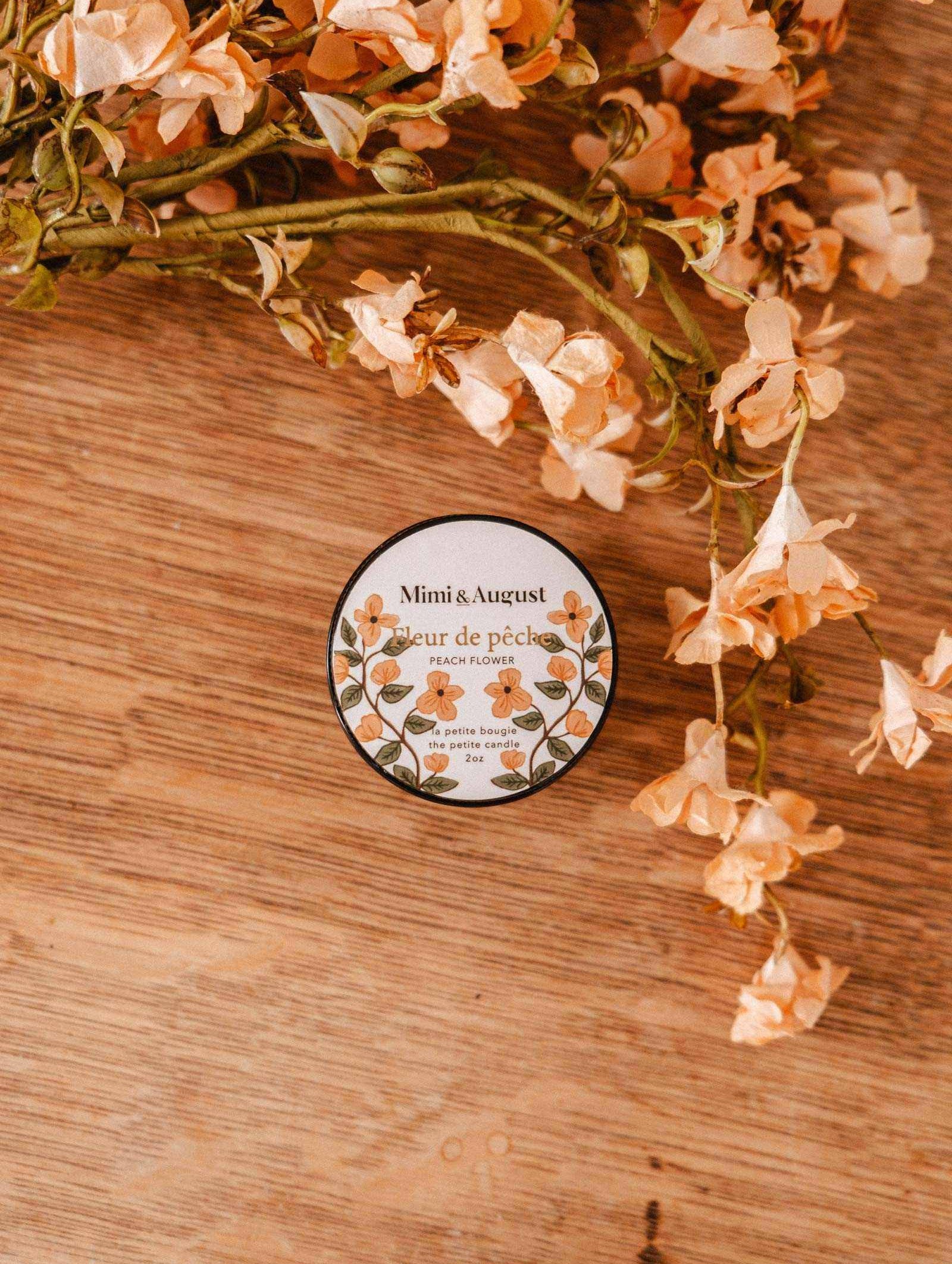 A 2 oz Mimi & August Fleur de Pêche soy candle rests on a wooden surface beside peach-toned artificial flowers, highlighting the hand-poured floral charm of this Montreal-made candle.