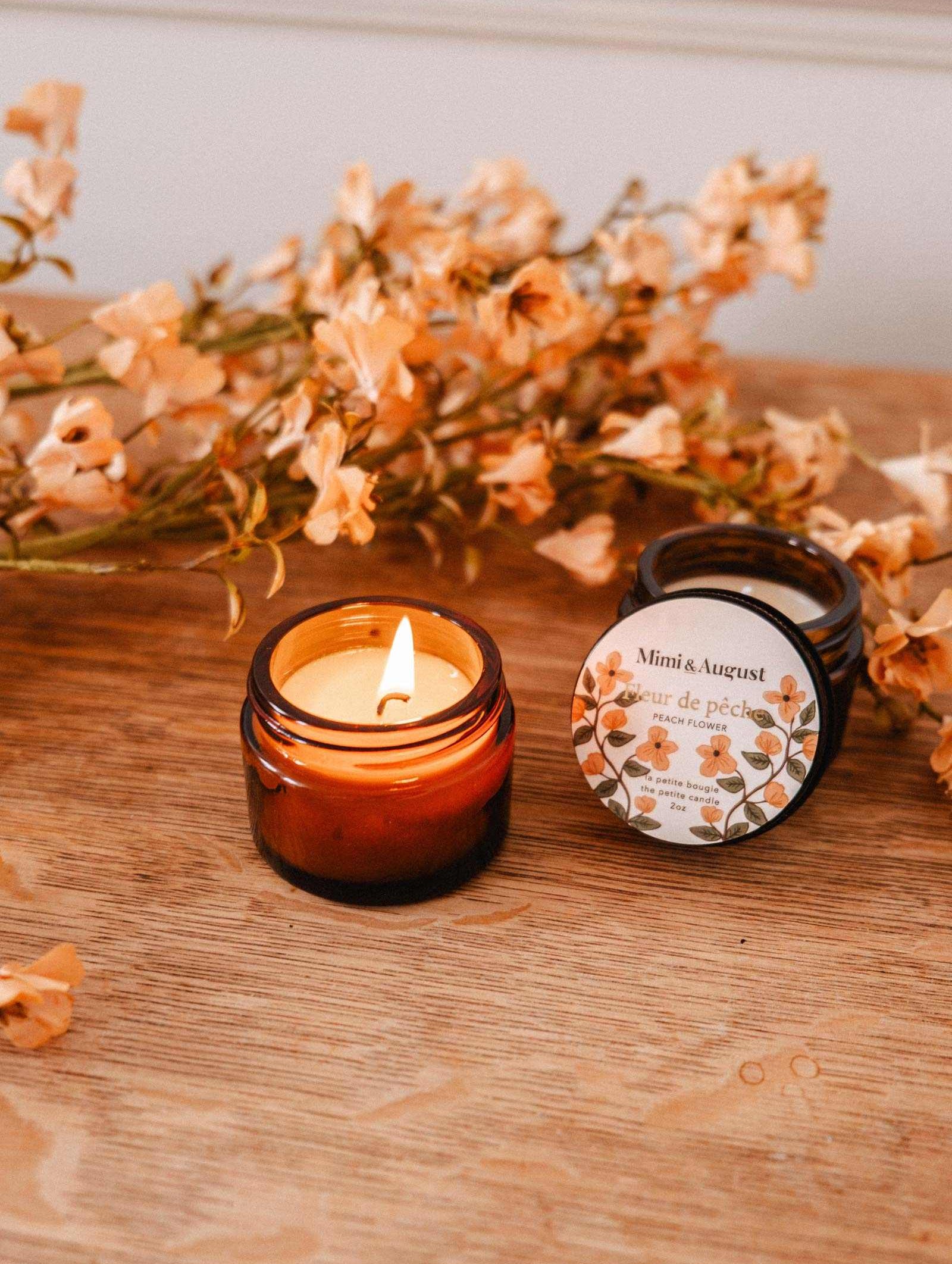 The Mimi & August Fleur de Pêche 2 oz soy candle, hand-poured in Montreal, glows in its amber glass jar on a wooden surface beside its lid, with peach-colored flowers in the background.
