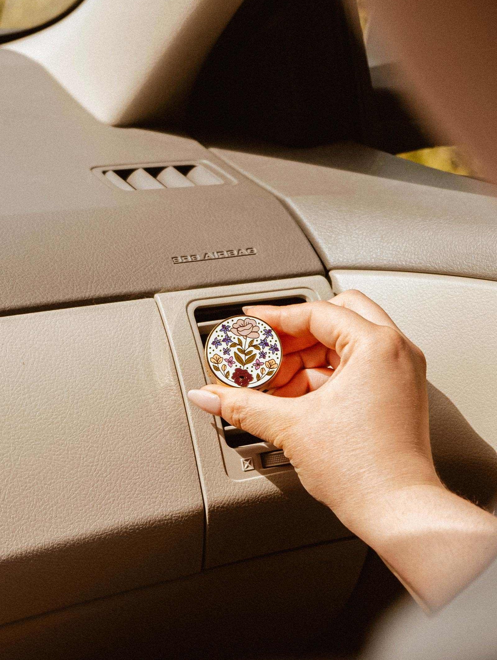 A hand places the Mimi & August Floral Car Diffuser, featuring a round floral design, into a car's air vent above a beige dashboard.