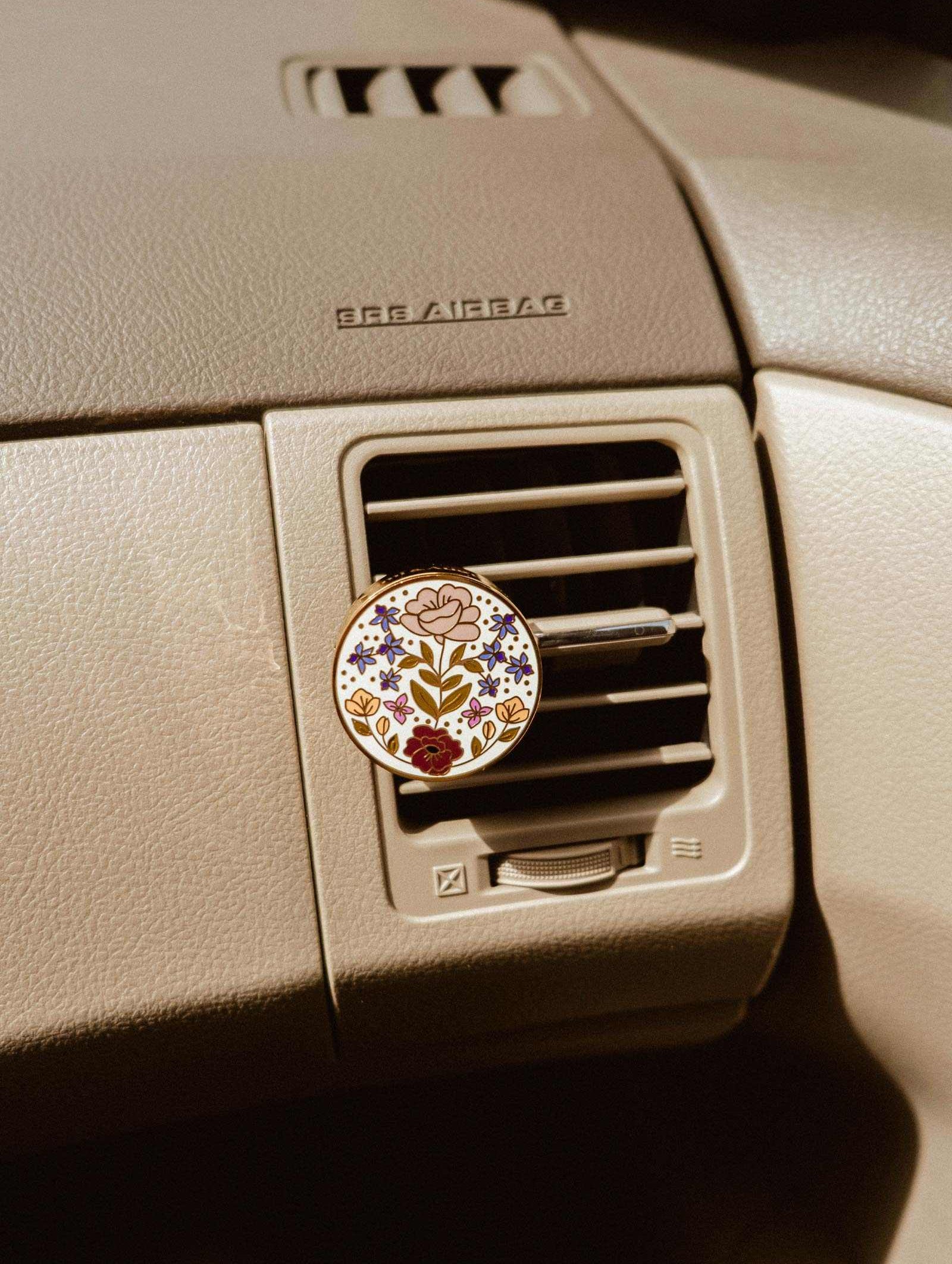 A Mimi & August Floral Car Diffuser is clipped to a car’s air vent on a beige dashboard labeled “SRS AIRBAG.”.