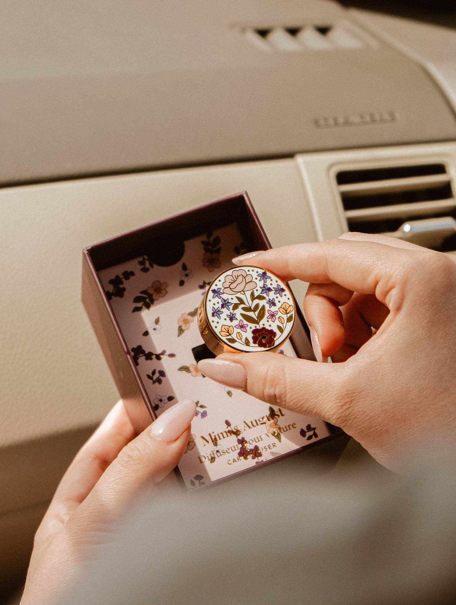 A person holds a Mimi & August Floral Car Diffuser tin above an open box with a matching floral design while sitting in a car.