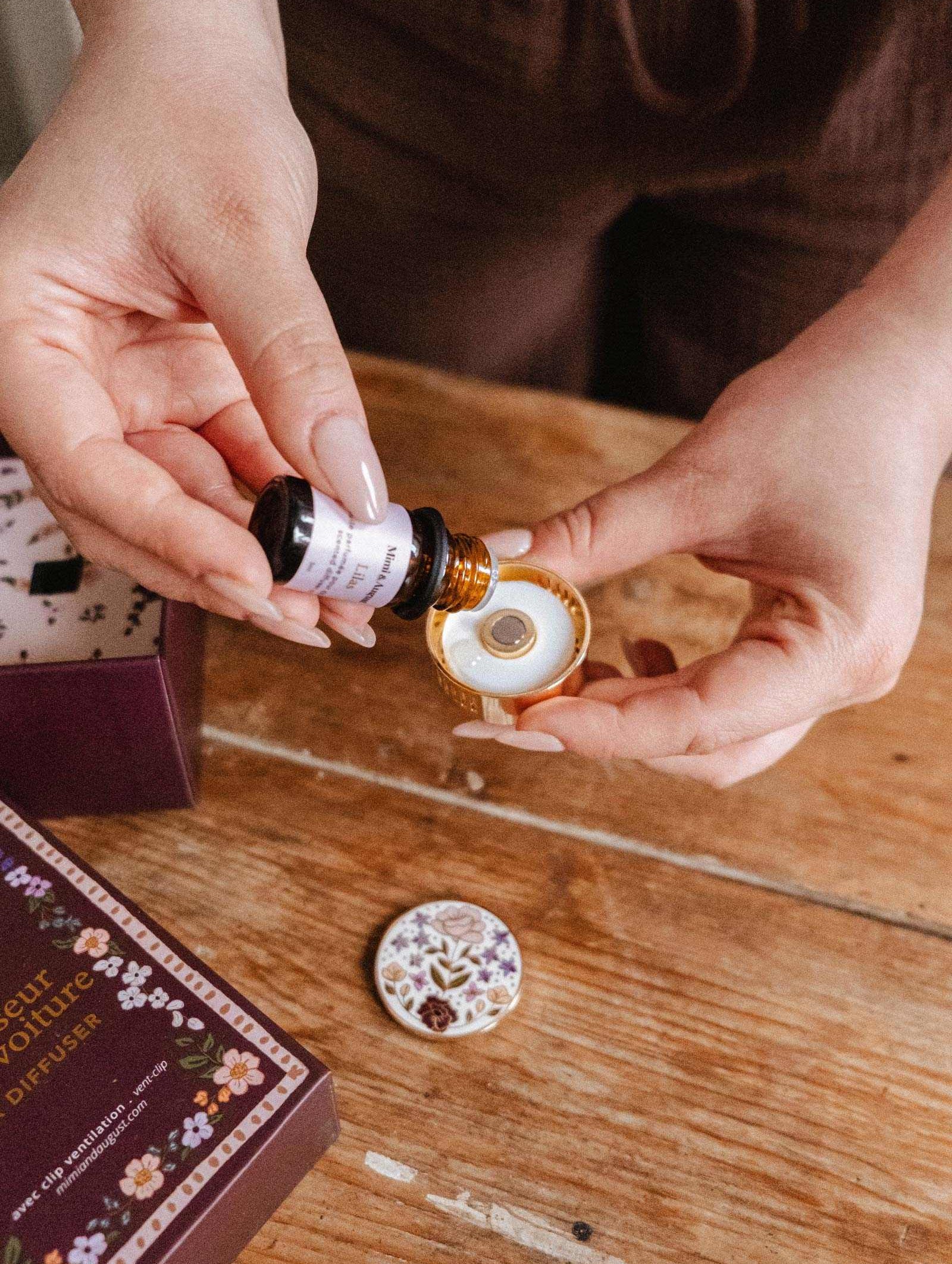 Someone pours liquid from a small brown bottle into the Mimi & August Floral Car Diffuser, a decorative round air freshener, on a wooden table beside its box and lid.