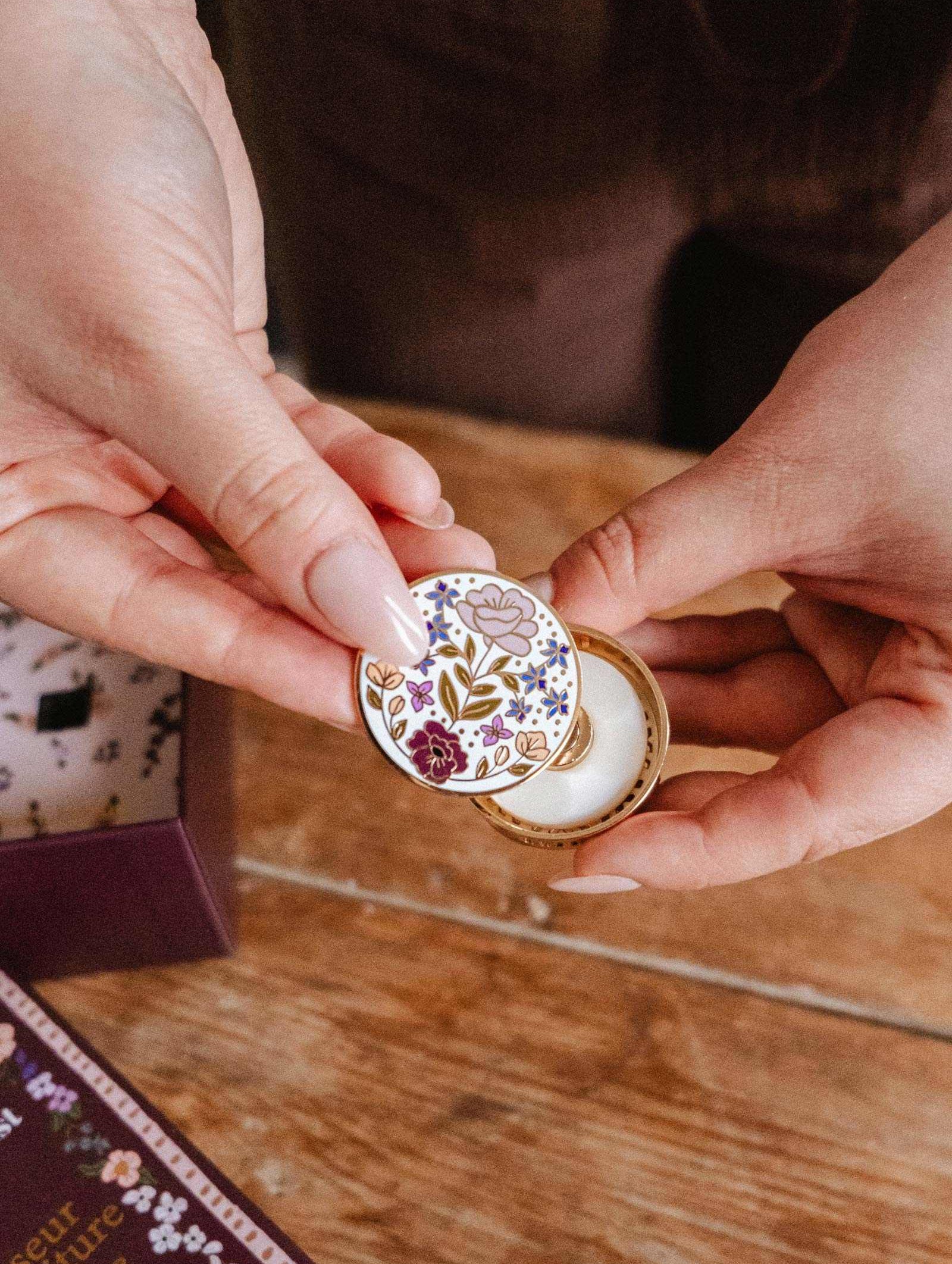 A person holds an open small round tin with a floral lid, revealing white balm inside—the Mimi & August Floral Car Diffuser that adds elegance and fragrance to your car.