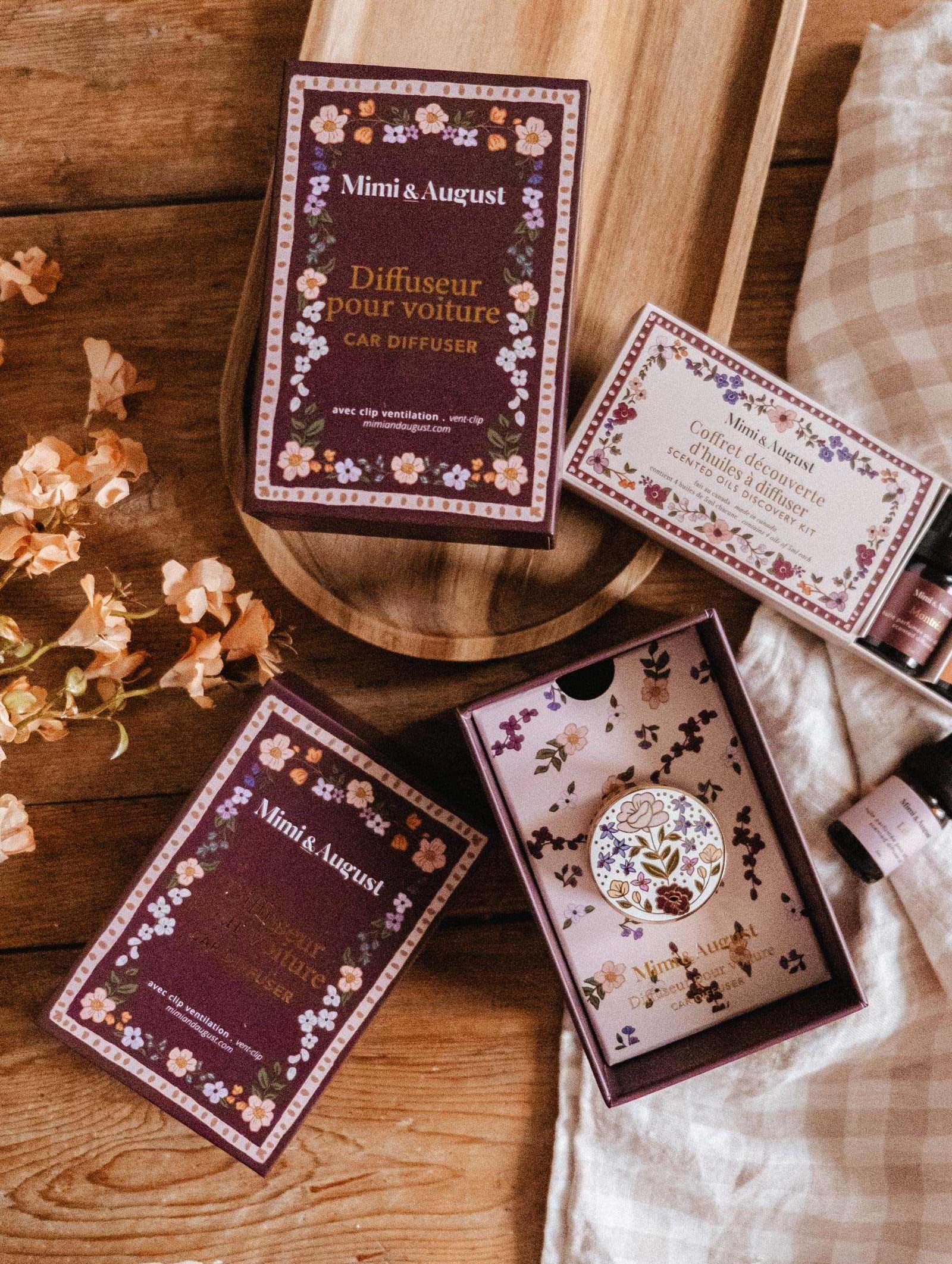 A wooden tray displays Mimi & August Floral Car Diffusers and scented candles, accented with small flowers, on a wooden table draped with a checkered cloth.