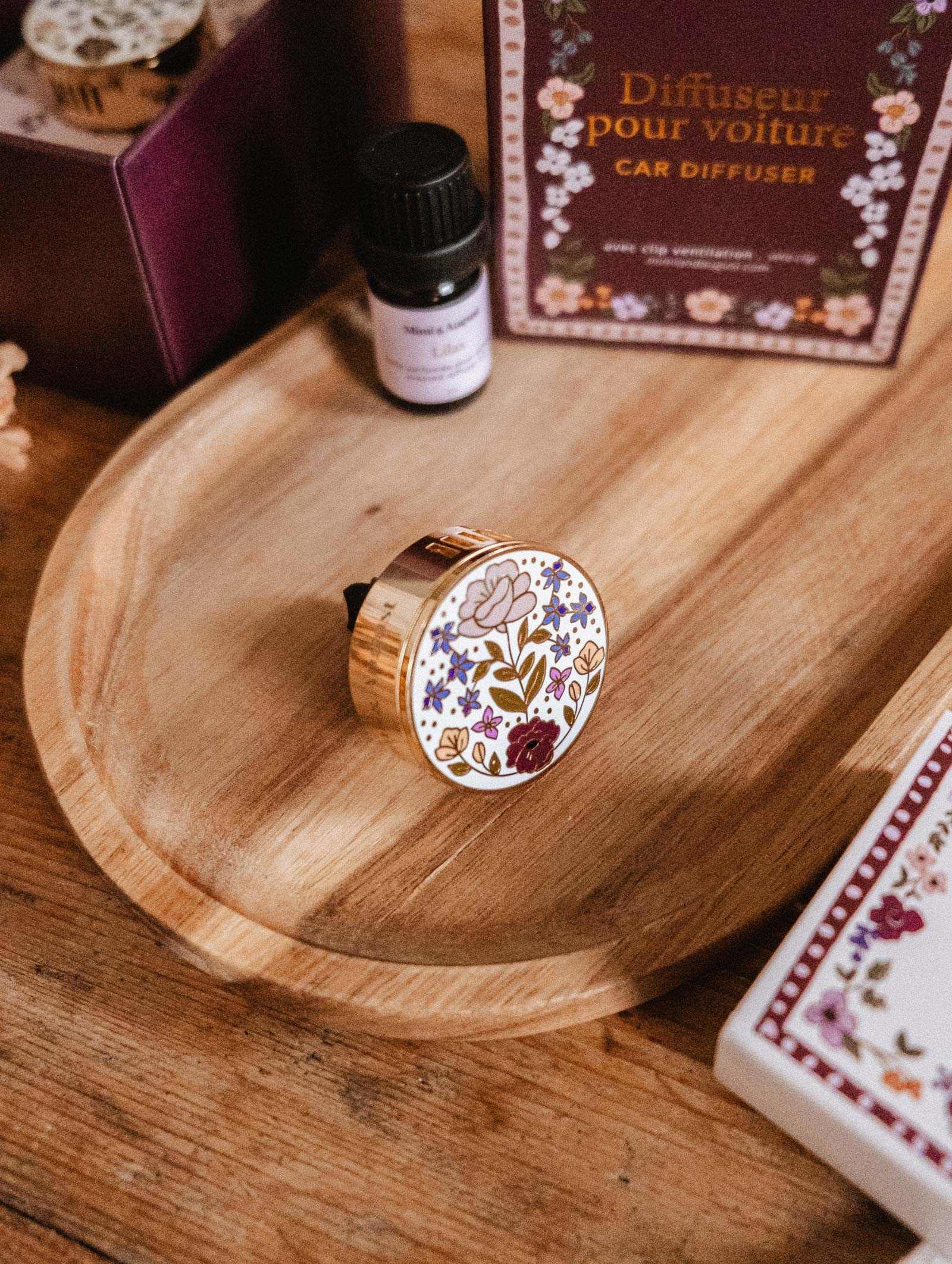 The Mimi & August Floral Car Diffuser, featuring a round floral design, is displayed on a wooden plate next to an essential oil bottle and a box labeled "Diffuser pour voiture.