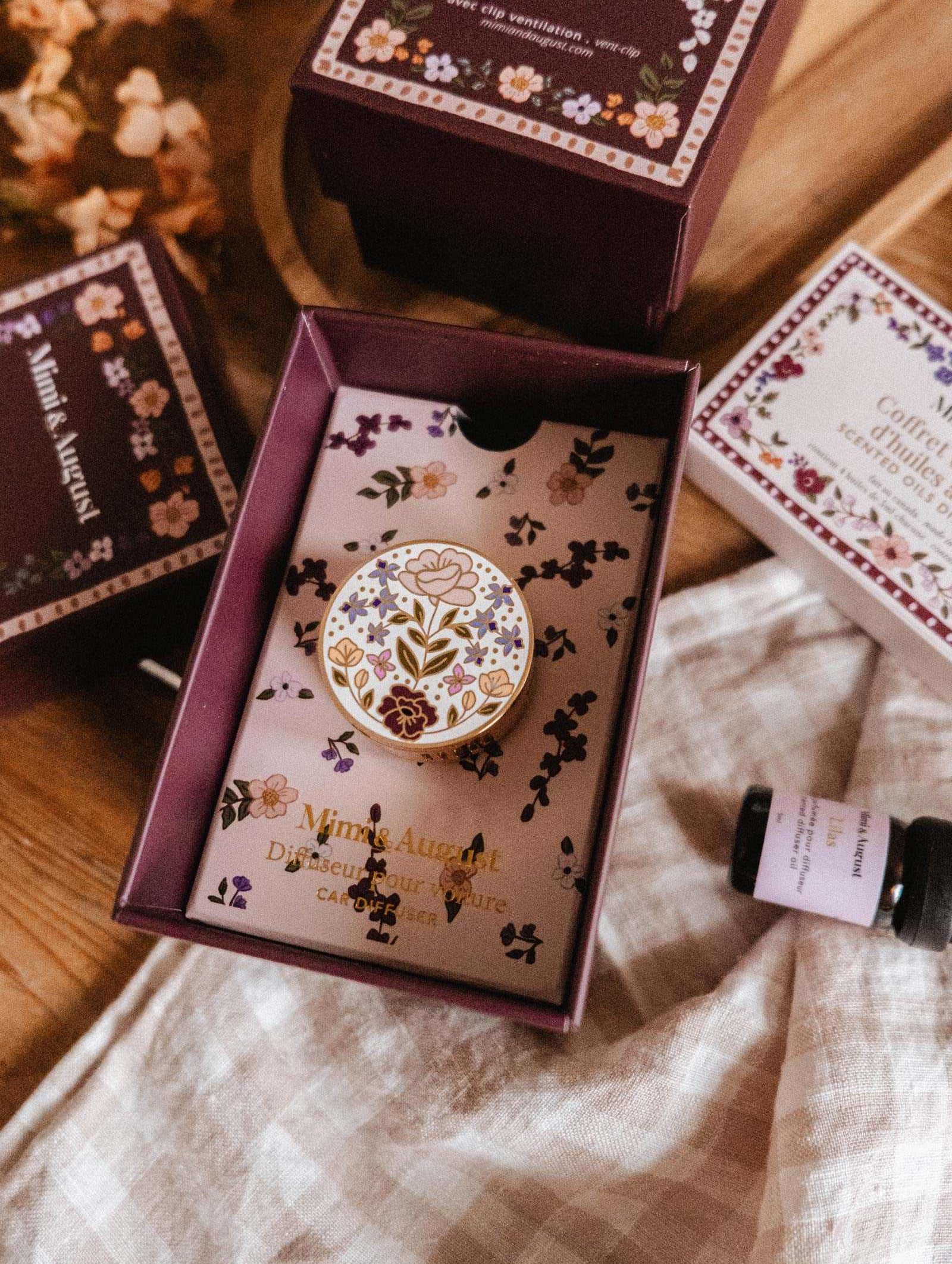 The Mimi & August Floral Car Diffuser is a decorative floral enamel pin, showcased in an open purple box among similar boxes and a glass vial on wood—ideal as an enamel car diffuser or unique car air freshener accent.